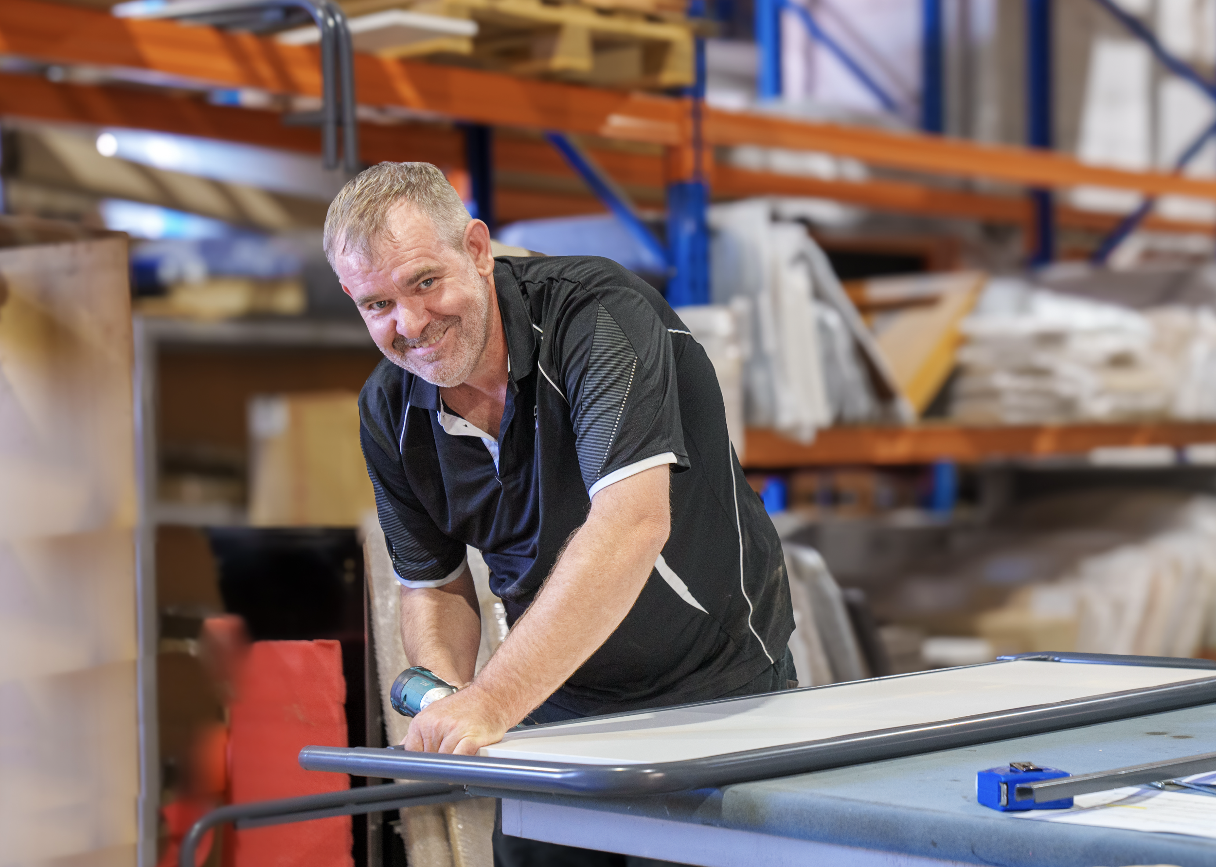 A man smiling and working with a power tool at a workbench in a warehouse or workshop, surrounded by shelves with various materials.