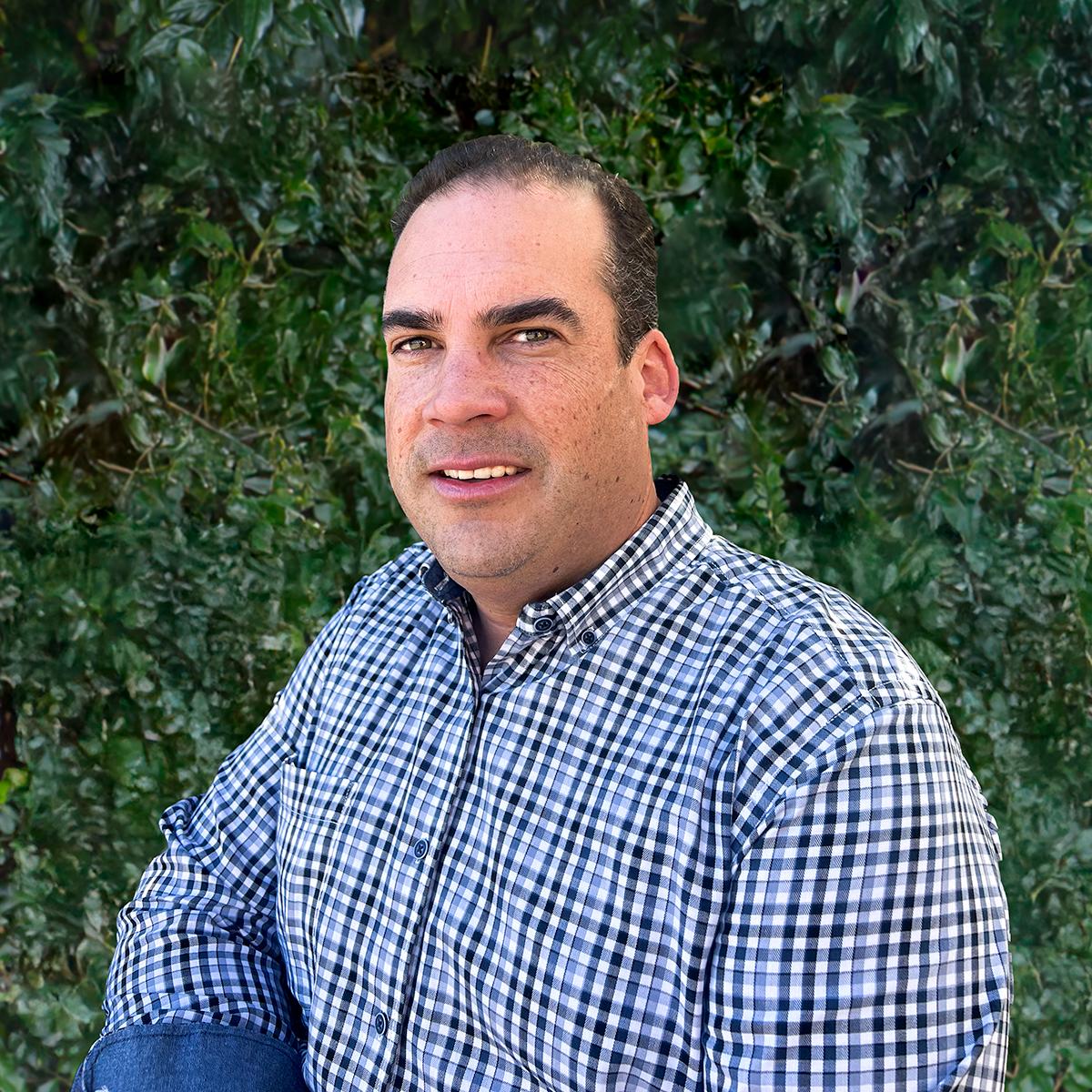 Man in checkered shirt standing outdoors in front of leafy green bushes.