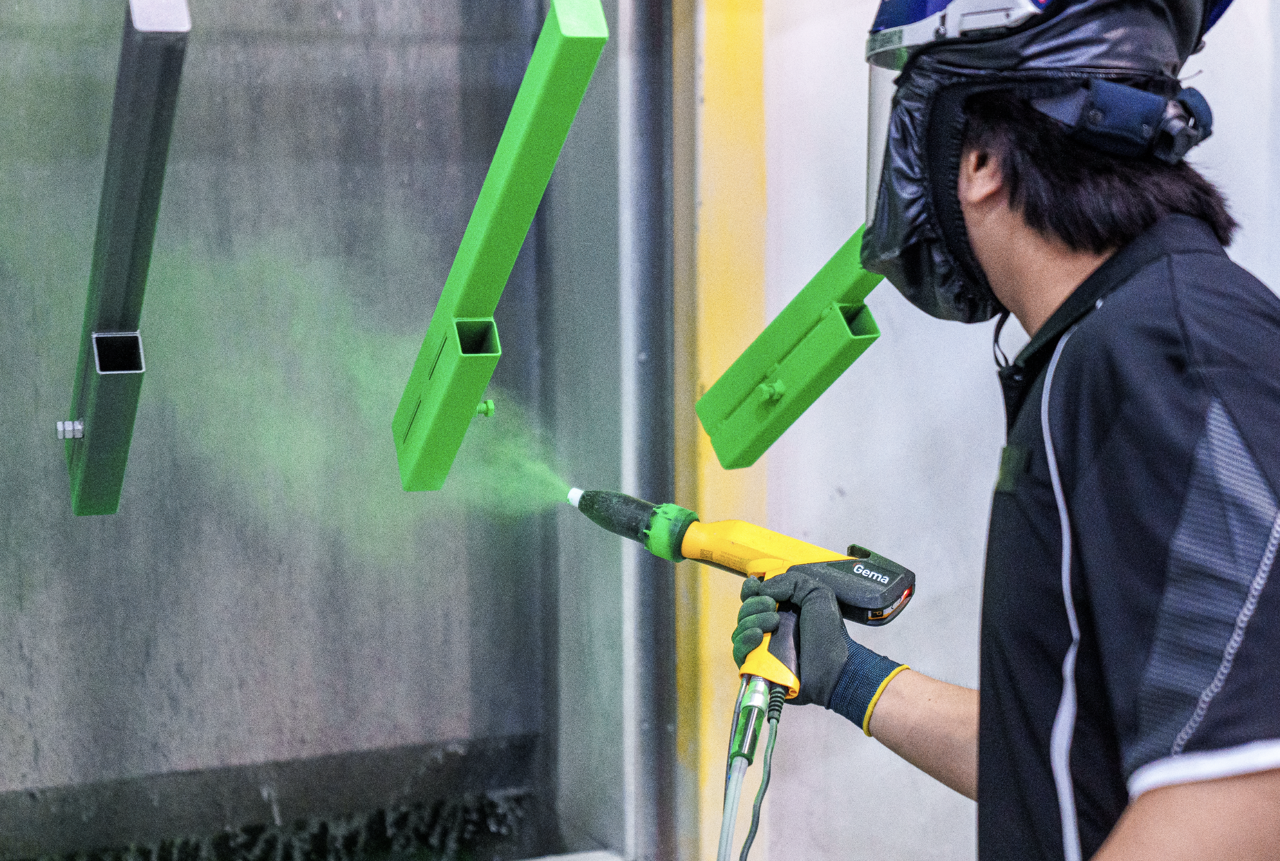 Worker wearing a black uniform, gloves, and safety gear, using a heat gun to attach or shape bright green plastic panels that are hanging on a metallic surface.