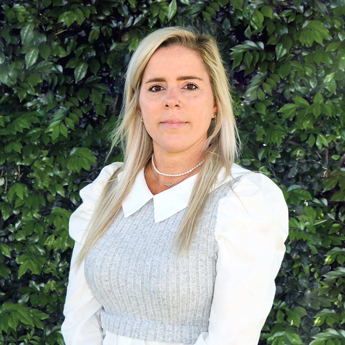 A woman with blonde hair, wearing a pearl necklace, gold earrings, and a white collared shirt under a gray sweater, standing in front of green foliage.