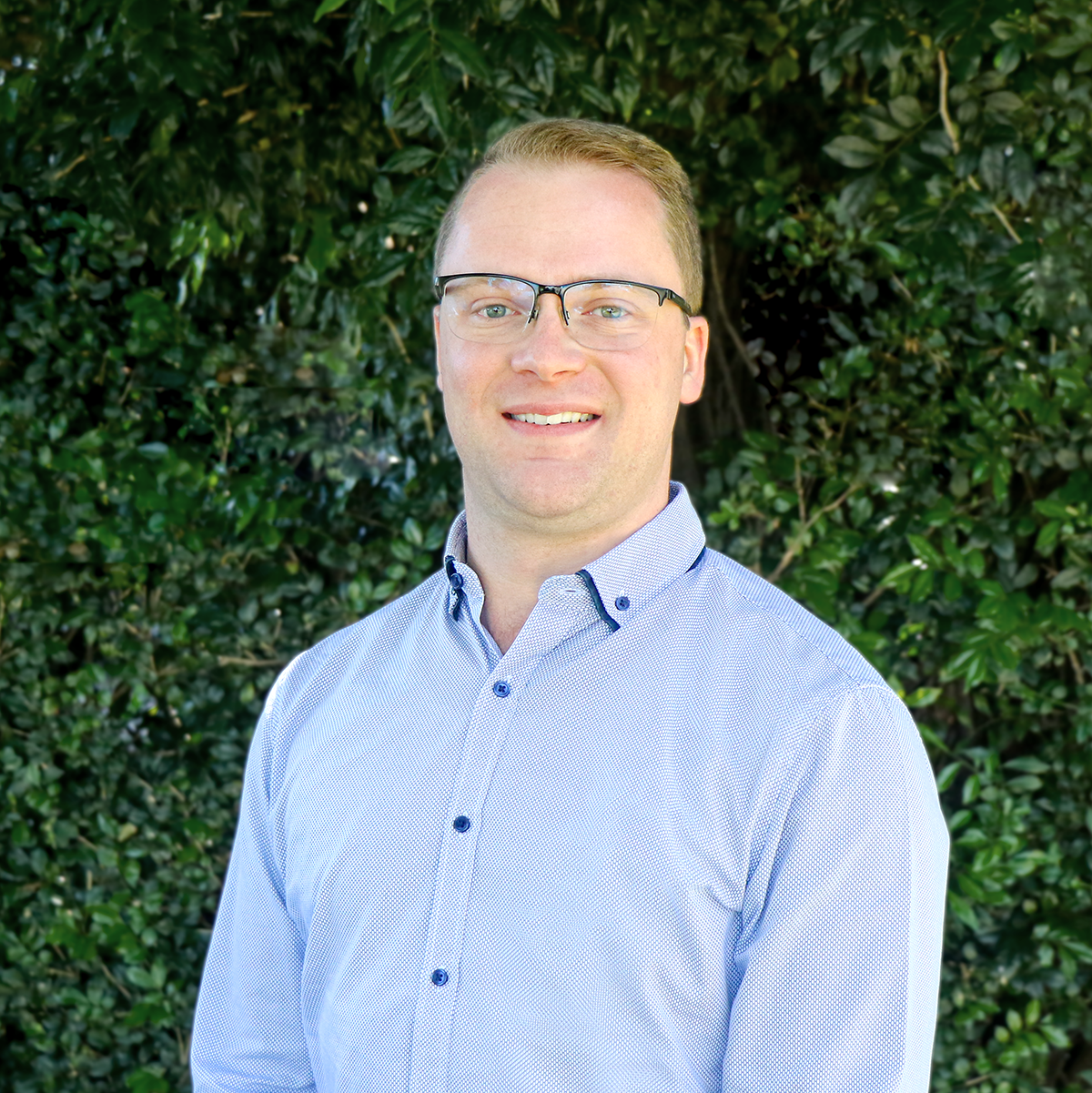 Portrait of a smiling man wearing glasses and a light blue button-up shirt, standing outdoors in front of green foliage.
