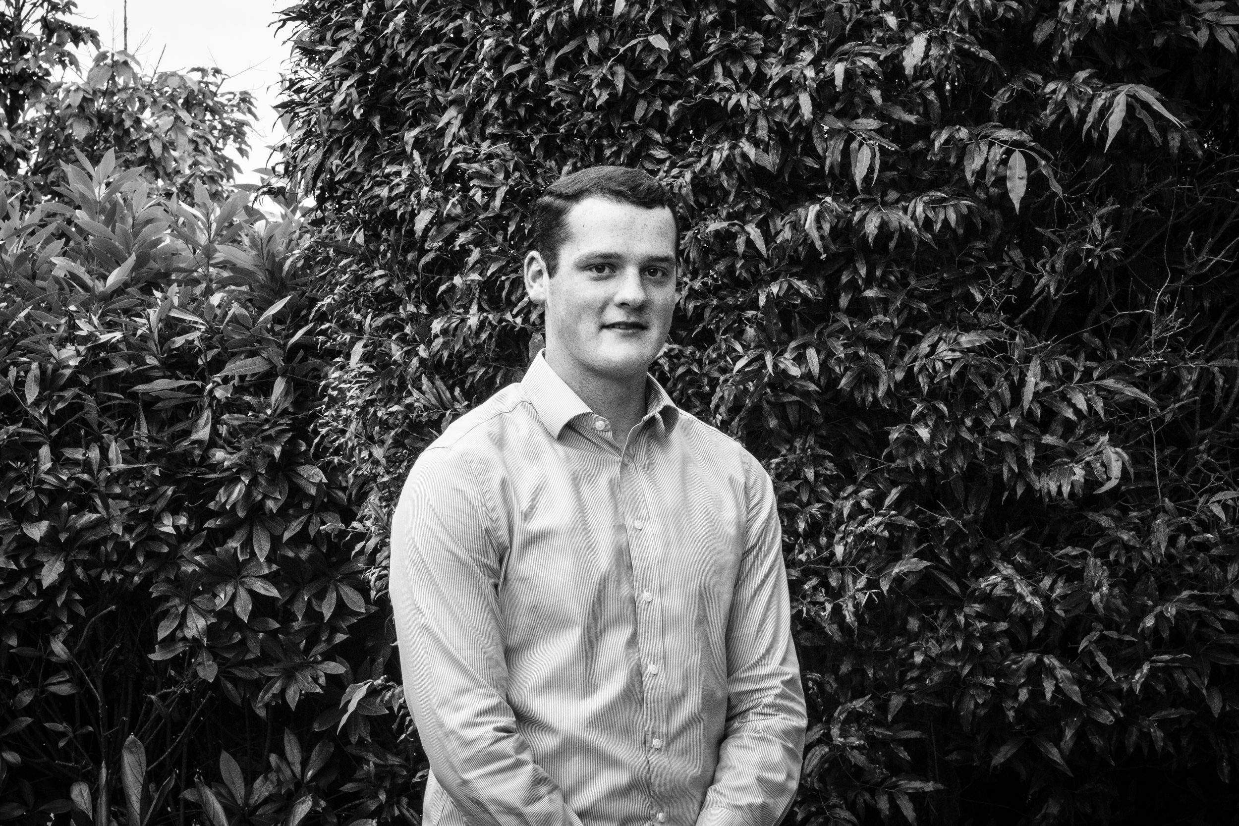 A young man standing outdoors in front of dense foliage, wearing a light-colored collared shirt and smiling slightly.
