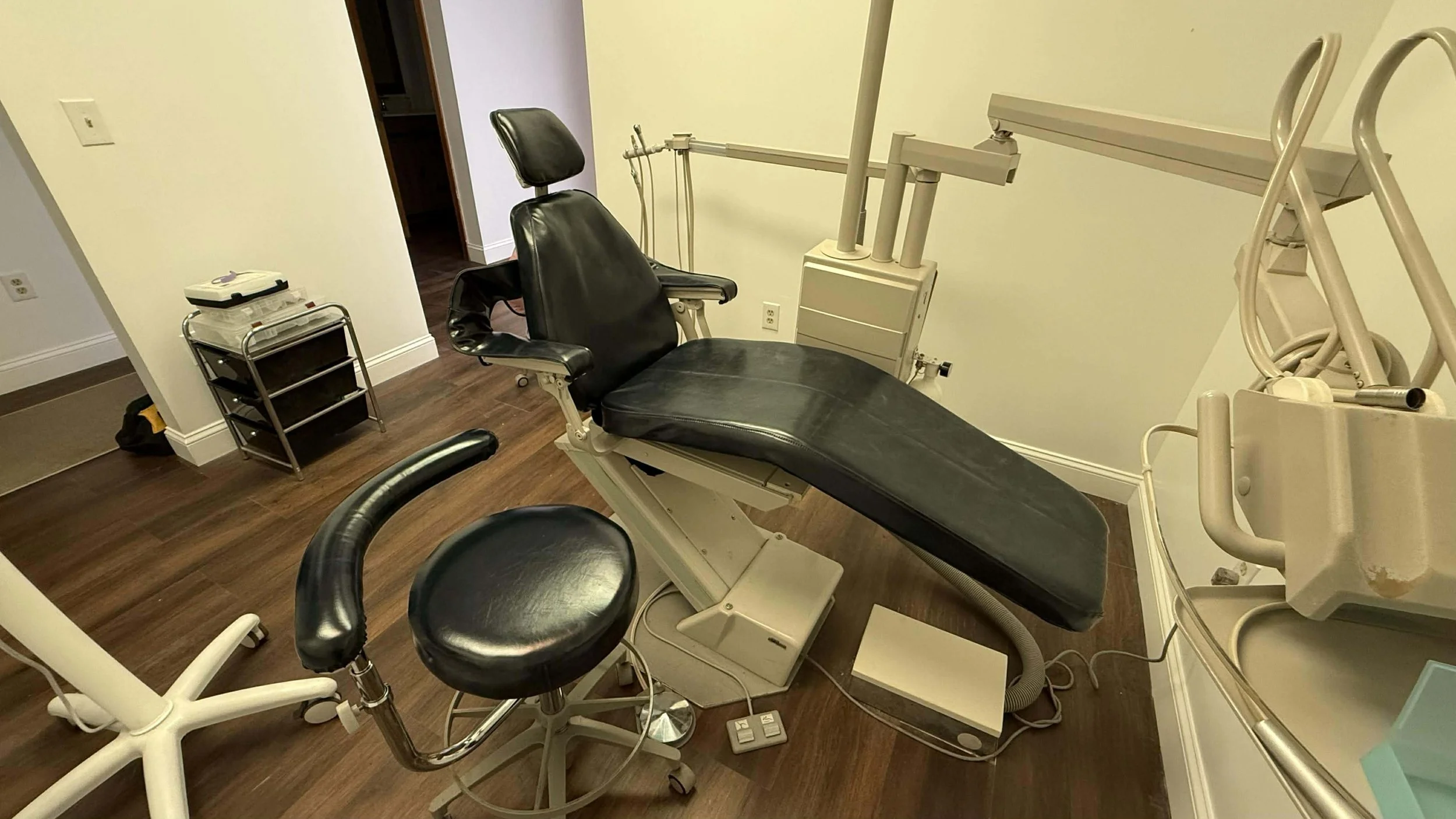 Medical examination room with a black examination chair, a black stool, medical equipment, and a small rolling cart against a white wall with wood flooring.