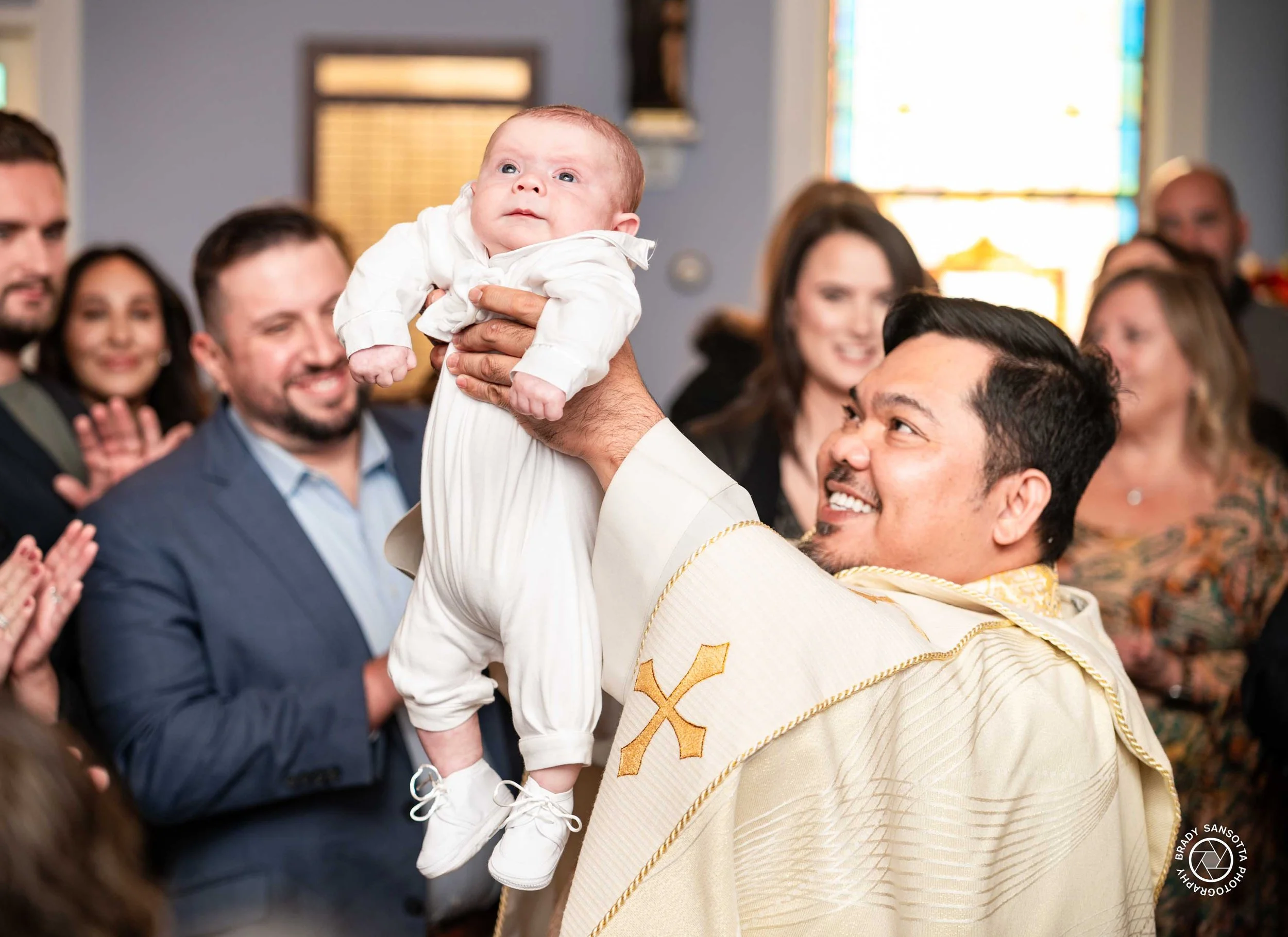 Priest lifting baby while family smiles during baptism ceremony at Immaculate Conception Church, Norwood, NJ.