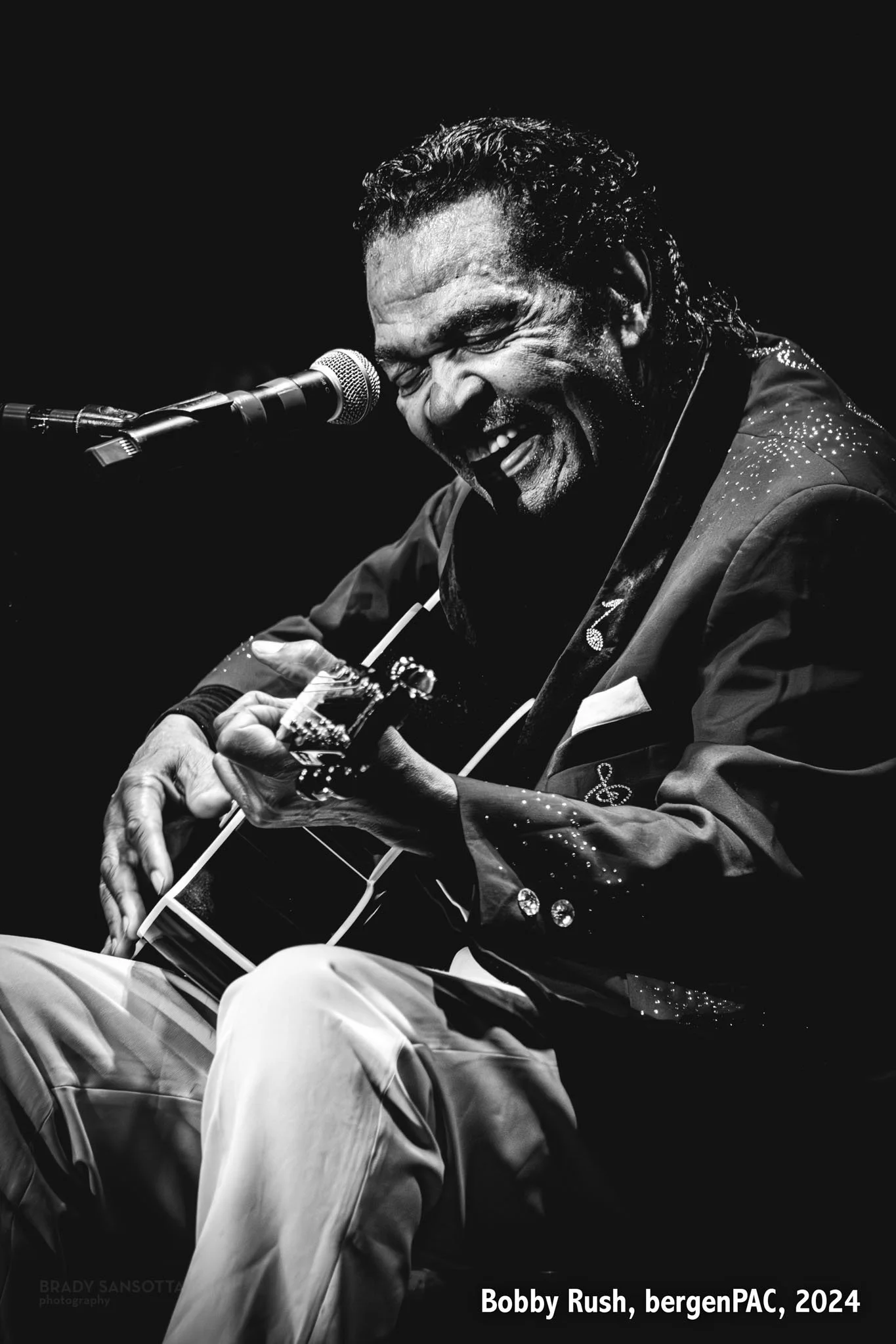 Black and white portrait of blues legend Bobby Rush smiling while playing guitar during a live show at BergenPAC in Englewood, NJ.