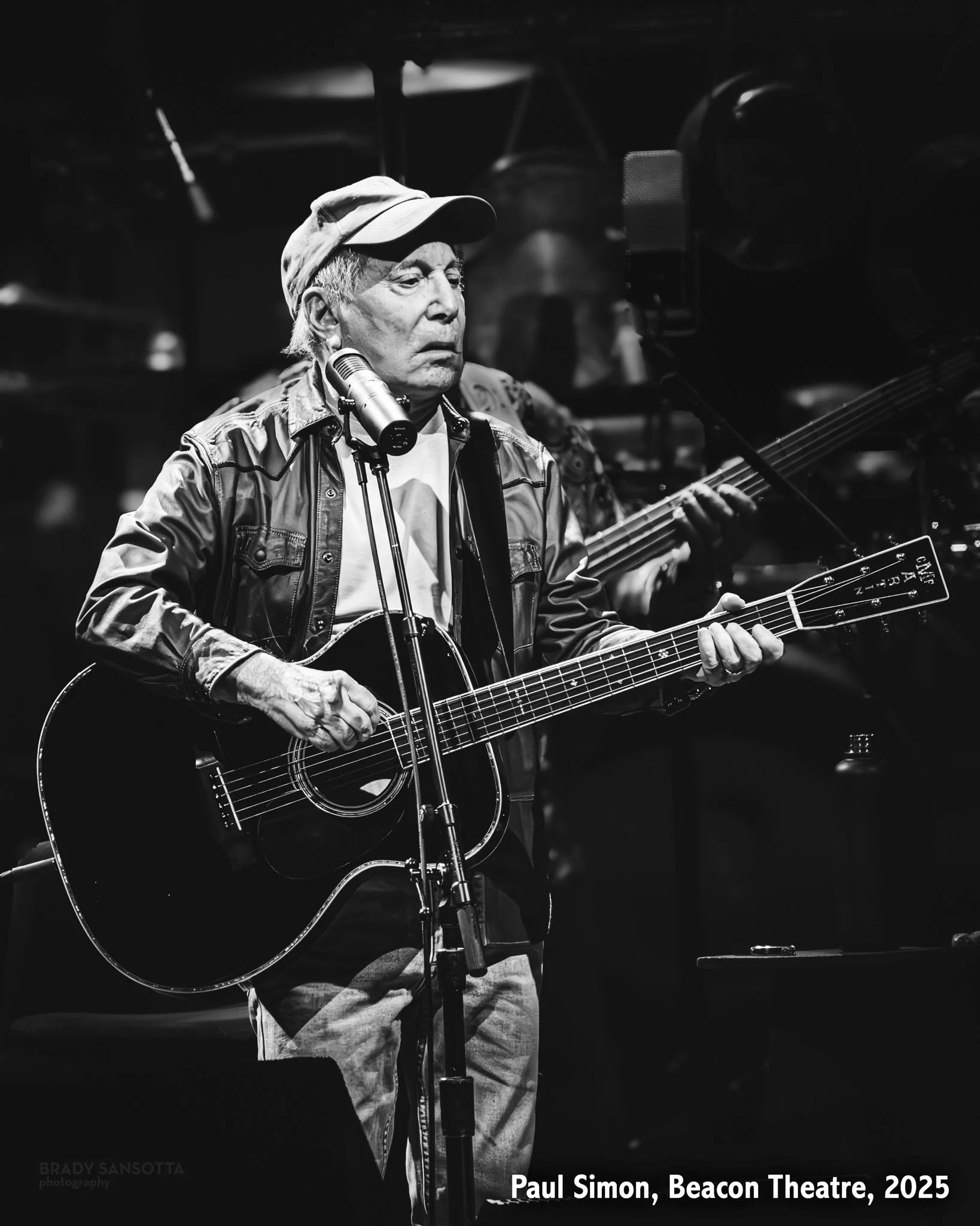Black and white photo of Paul Simon playing acoustic guitar and singing at his "A Quiet Celebration Tour" at the Beacon Theatre.