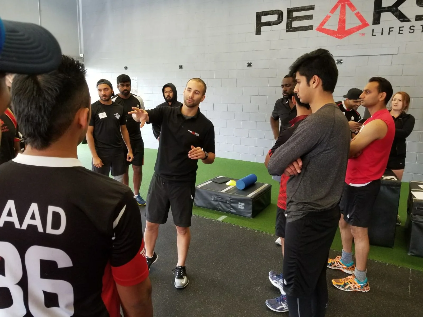 A trainer giving instructions to a group of people in a fitness training session at Peak Race gym, with various workout equipment around.