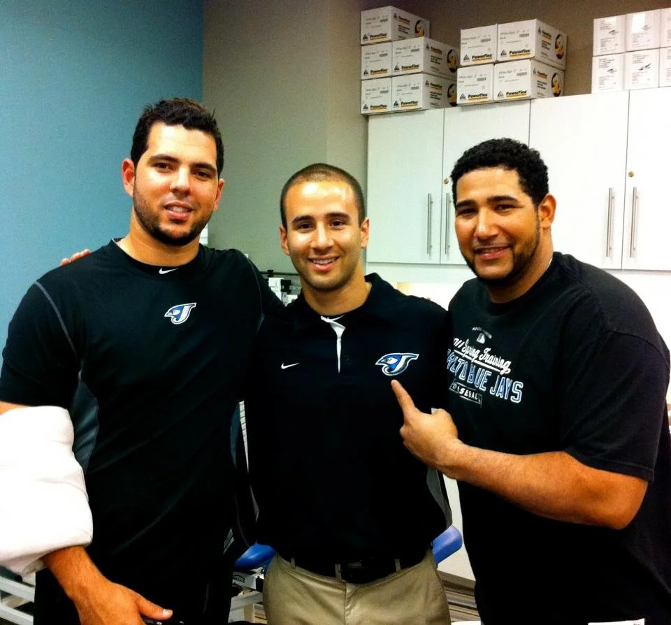 Three men standing together indoors, smiling at the camera, with medical supplies and cabinets in the background.