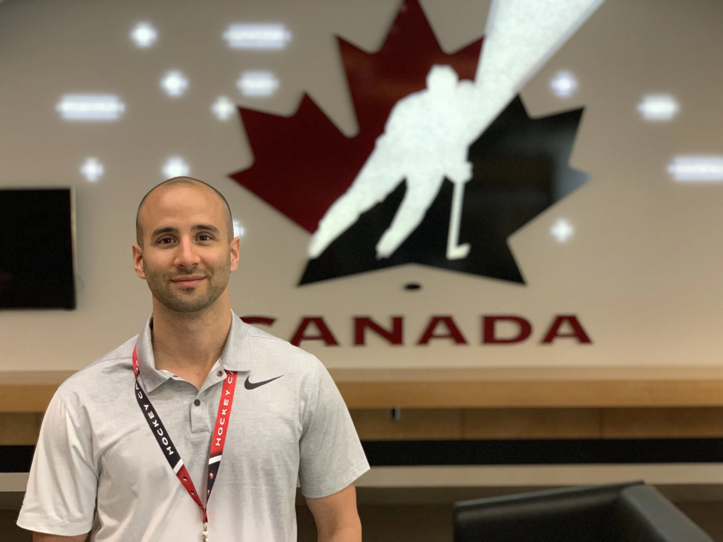 A man with a shaved head wearing a light gray Nike polo shirt and red and black hockey lanyard, standing in front of a Canadian hockey logo on the wall.