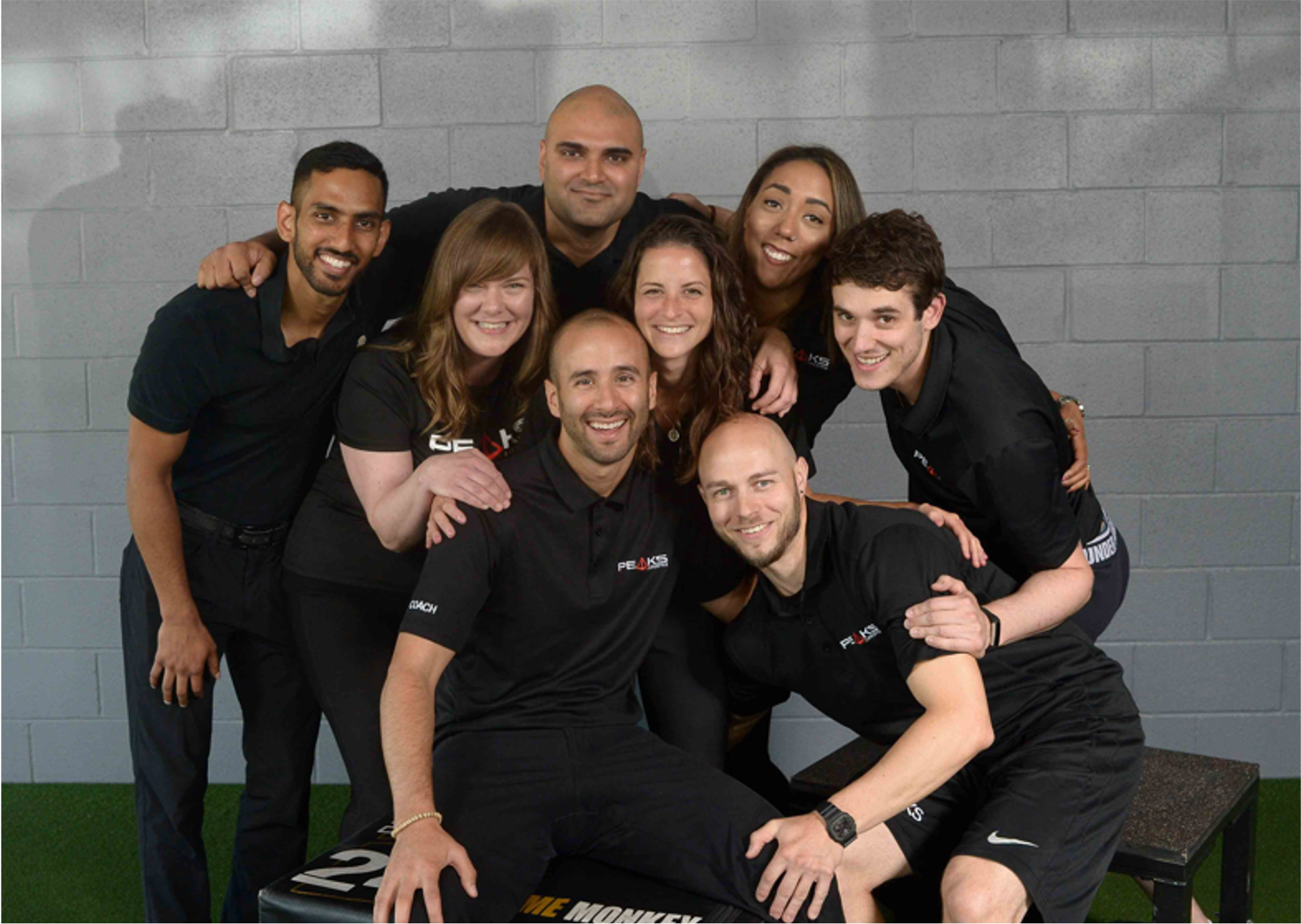 Group of nine diverse people smiling and posing together indoors against a gray brick wall.