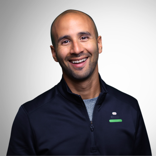 A smiling man with a bald head and beard wearing a black zip-up jacket against a plain light background.