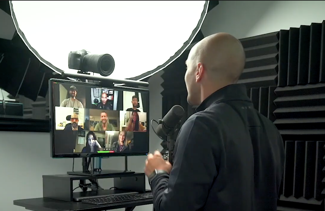 A man with a shaved head hosting a virtual meeting or podcast, speaking into a microphone in a soundproof studio, with a computer monitor showing multiple participants in a video call.