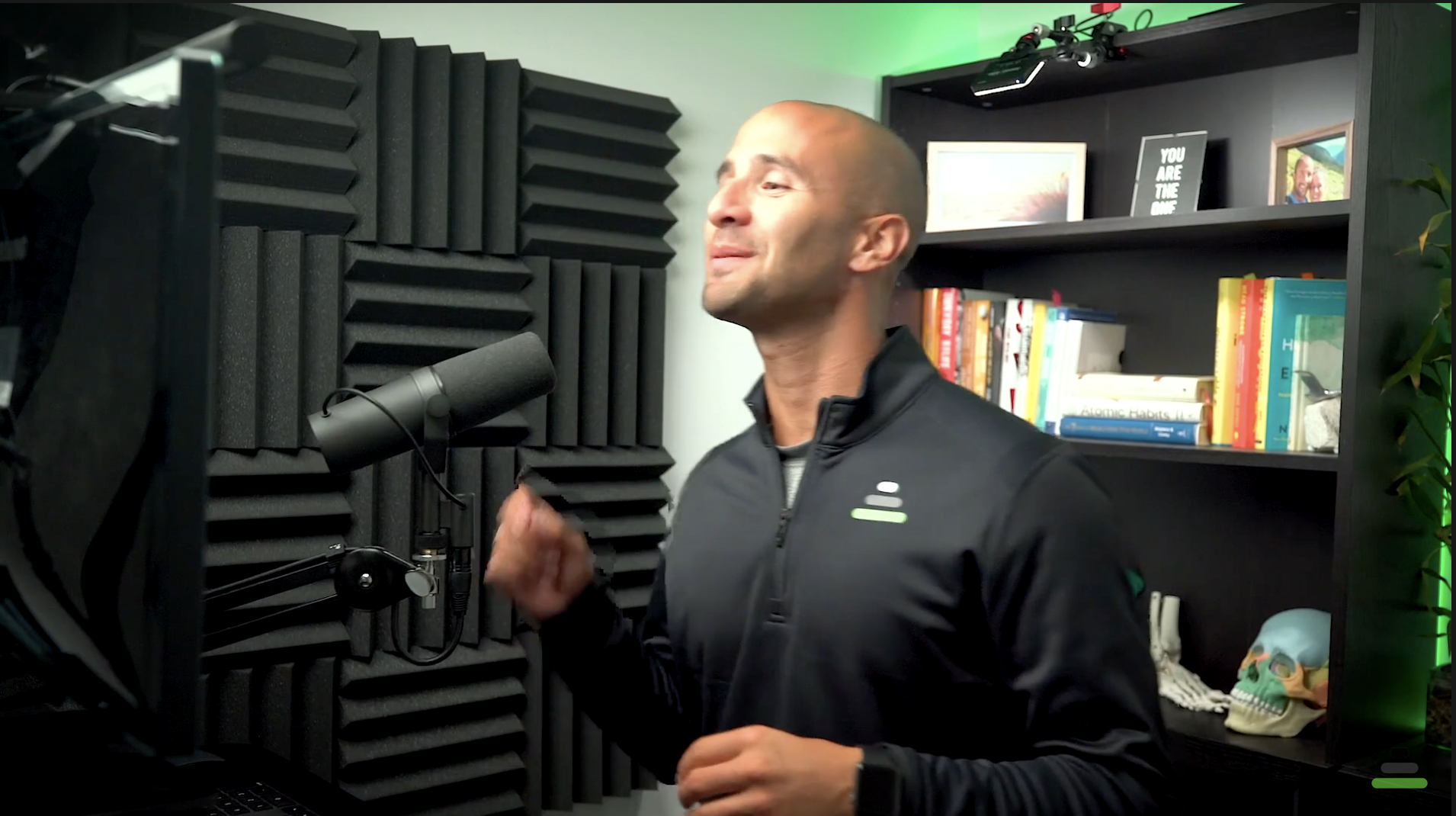 A man speaking into a microphone in a recording studio with black soundproof foam panels on the wall. There is a bookshelf with books, framed photos, and a model airplane behind him.