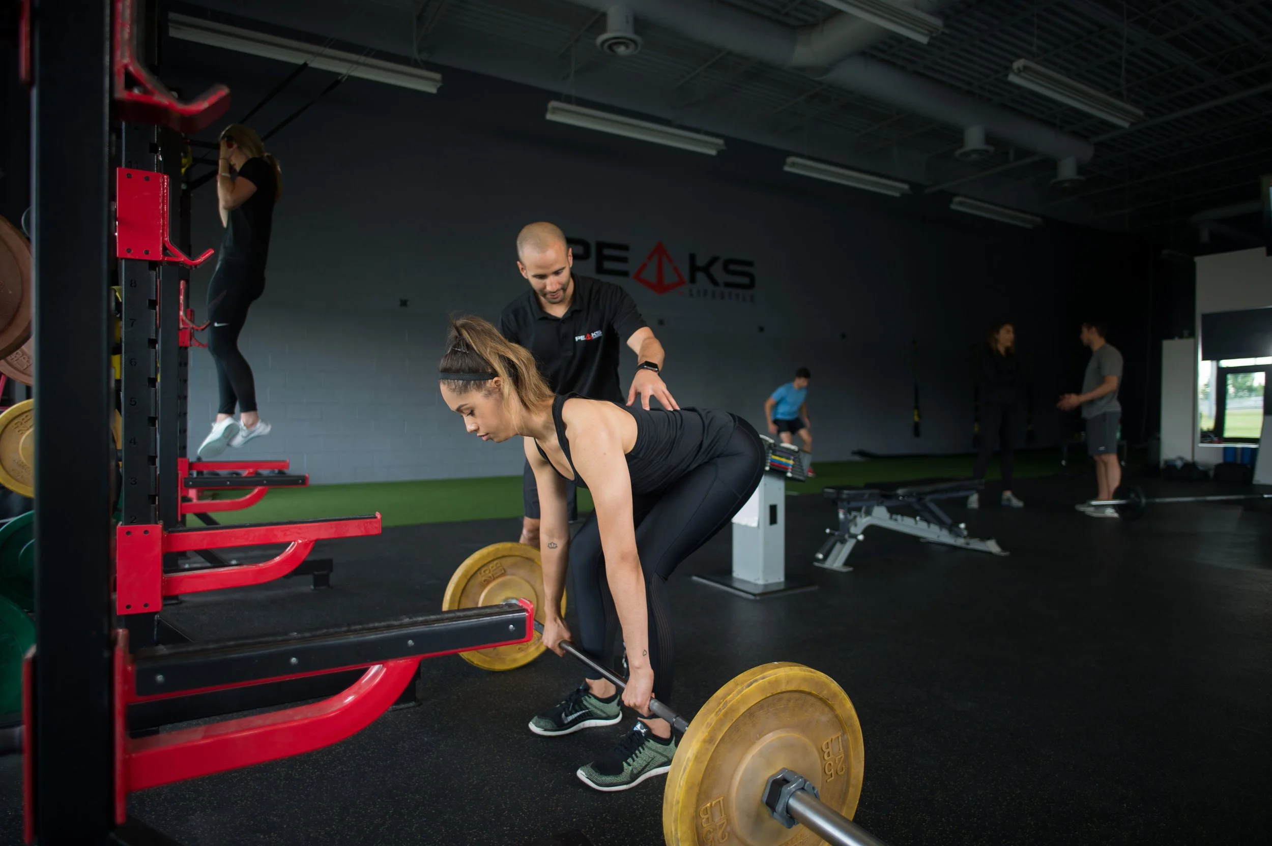 A woman lifting a barbell with yellow weights in a gym while a trainer observes. In the background, other people are working out and a woman is hanging from a pull-up bar.