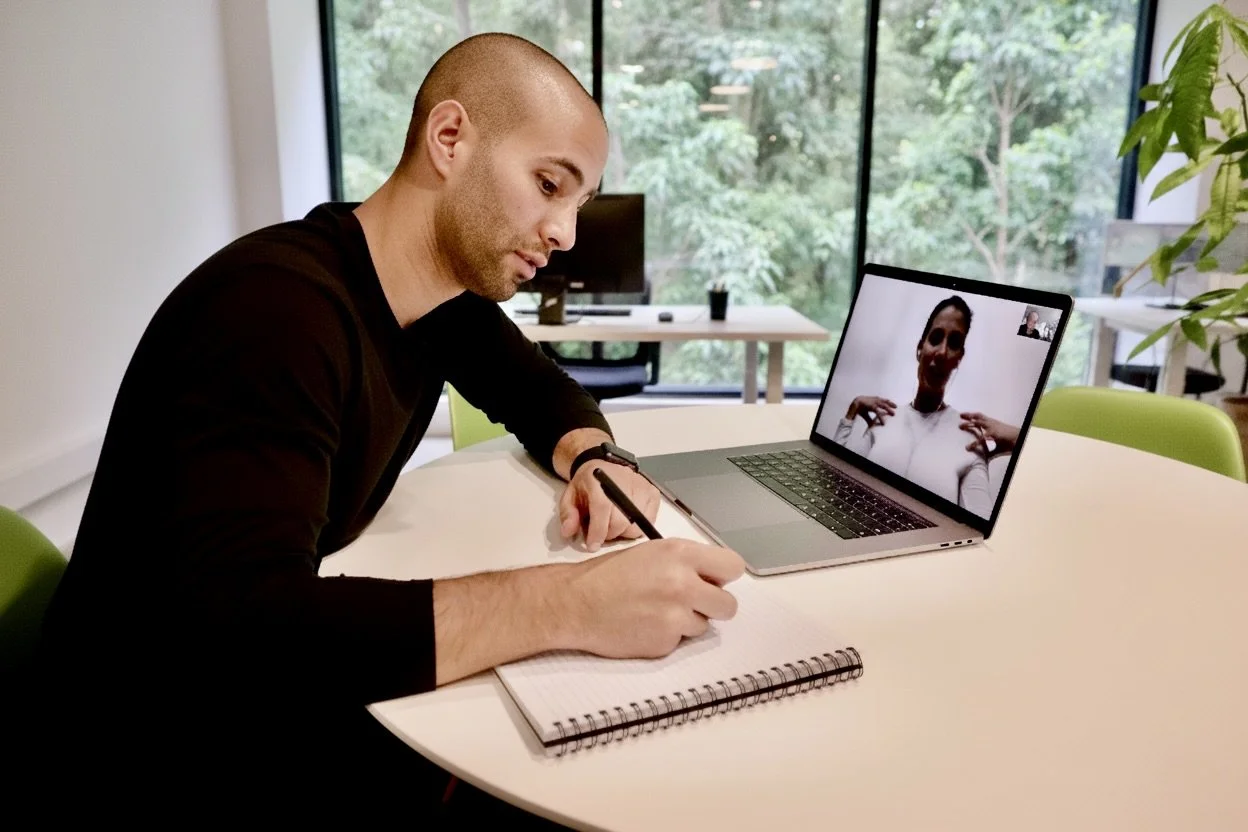 A man with a shaved head and black shirt sitting at a white table, taking notes in a spiral notebook while on a video call on his laptop, with a woman smiling on the screen.