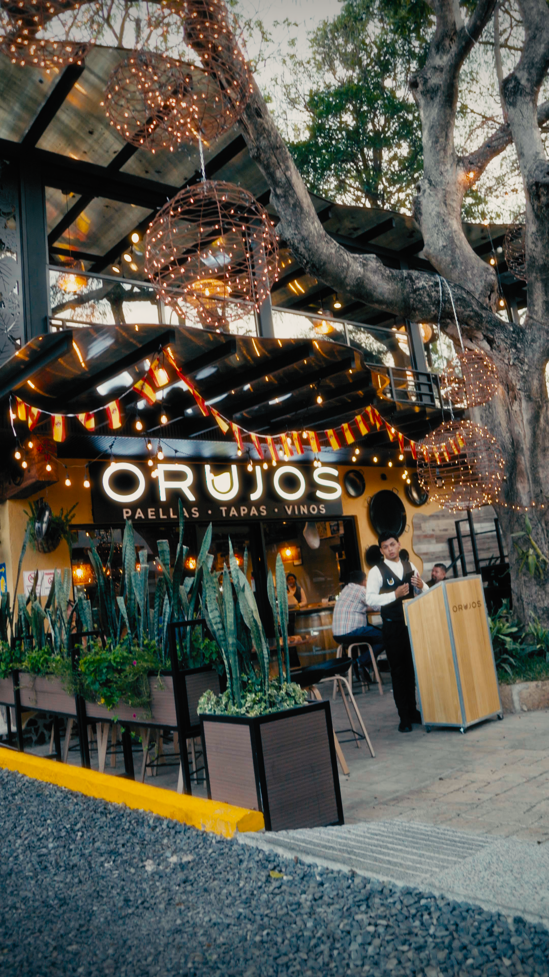 Exterior of a restaurant named 'Orujos' with string lights, hanging wire lanterns, and potted plants outside. A server stands near the entrance, and some patrons are seated inside visible through the window.