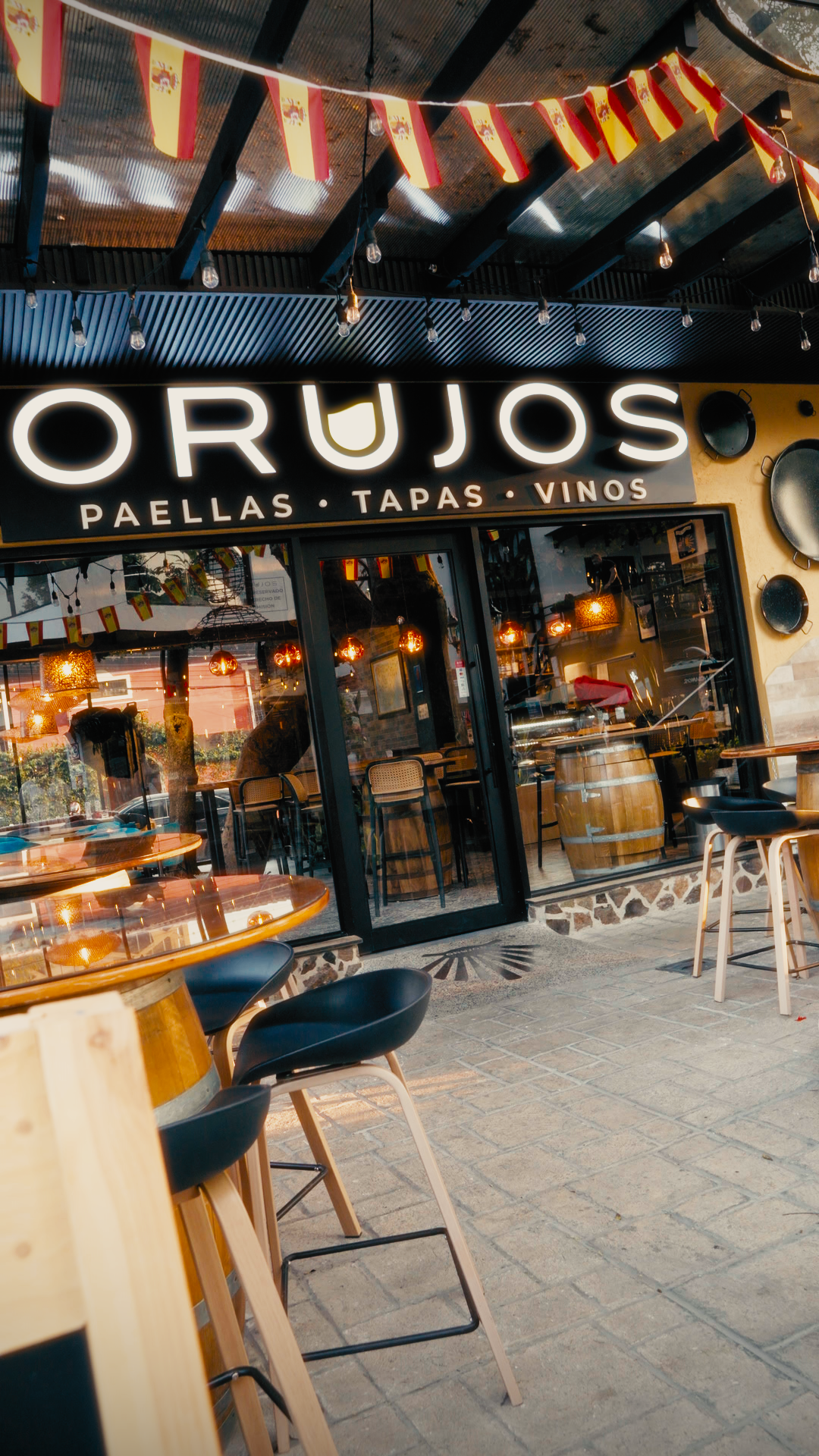 Exterior of a restaurant called 'Orujos' with a large illuminated sign, outdoor seating with black and wooden chairs, flags and string lights decorating the area, reflecting in the glass doors.
