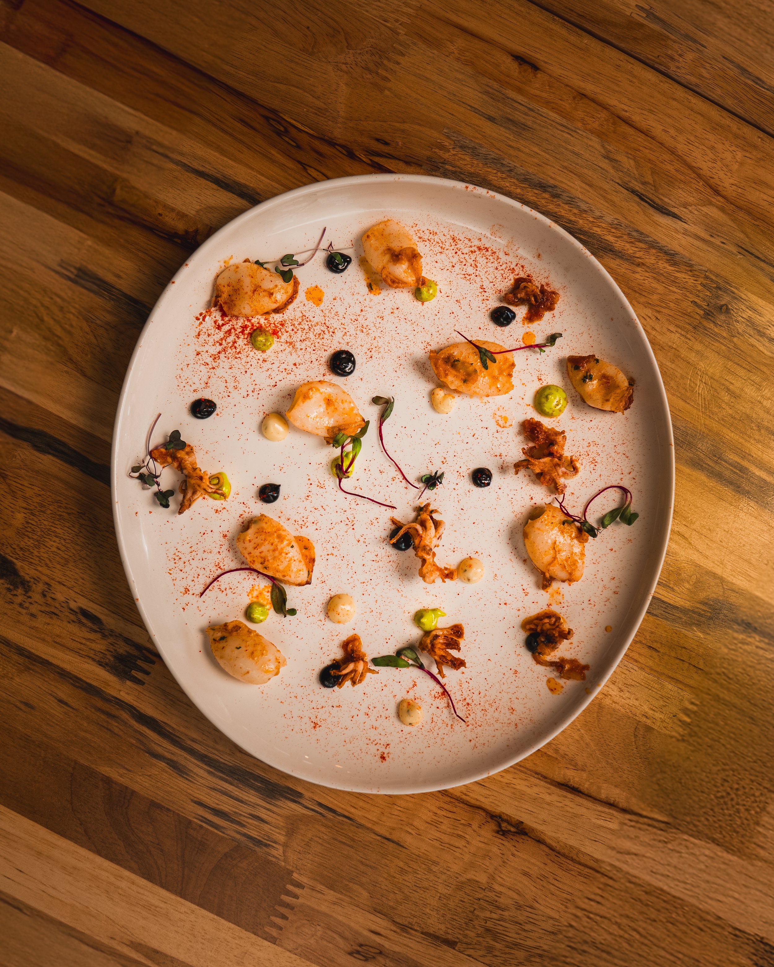 A white plate with small portions of food, garnished with microgreens, black and green dots, and a dusting of red spice, placed on a wooden surface.