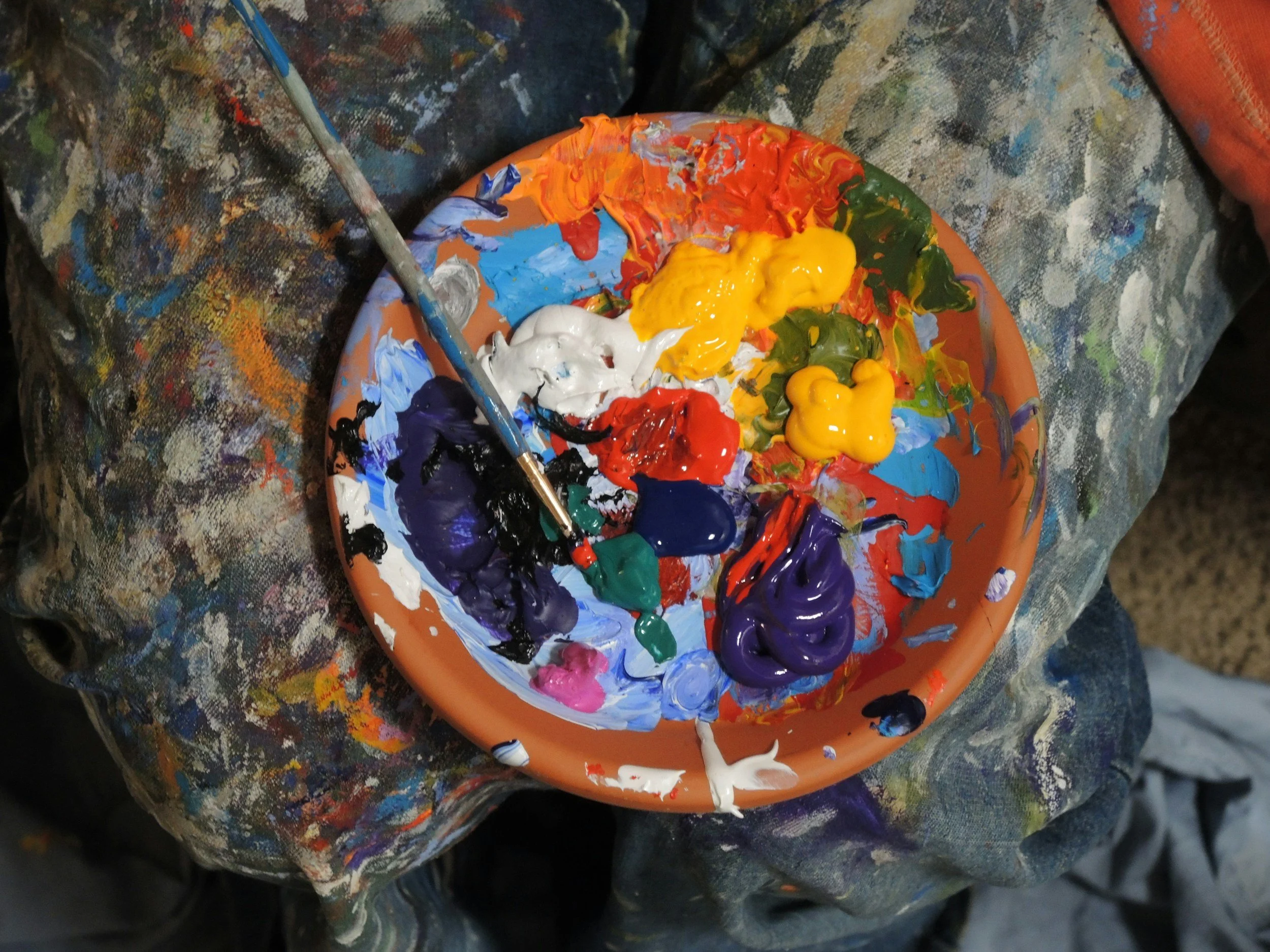A paint palette with various colors of paint, including white, black, yellow, red, blue, green, purple, and pink, resting on a heavily splattered and stained painter's cloth. A paintbrush is also resting on the palette.