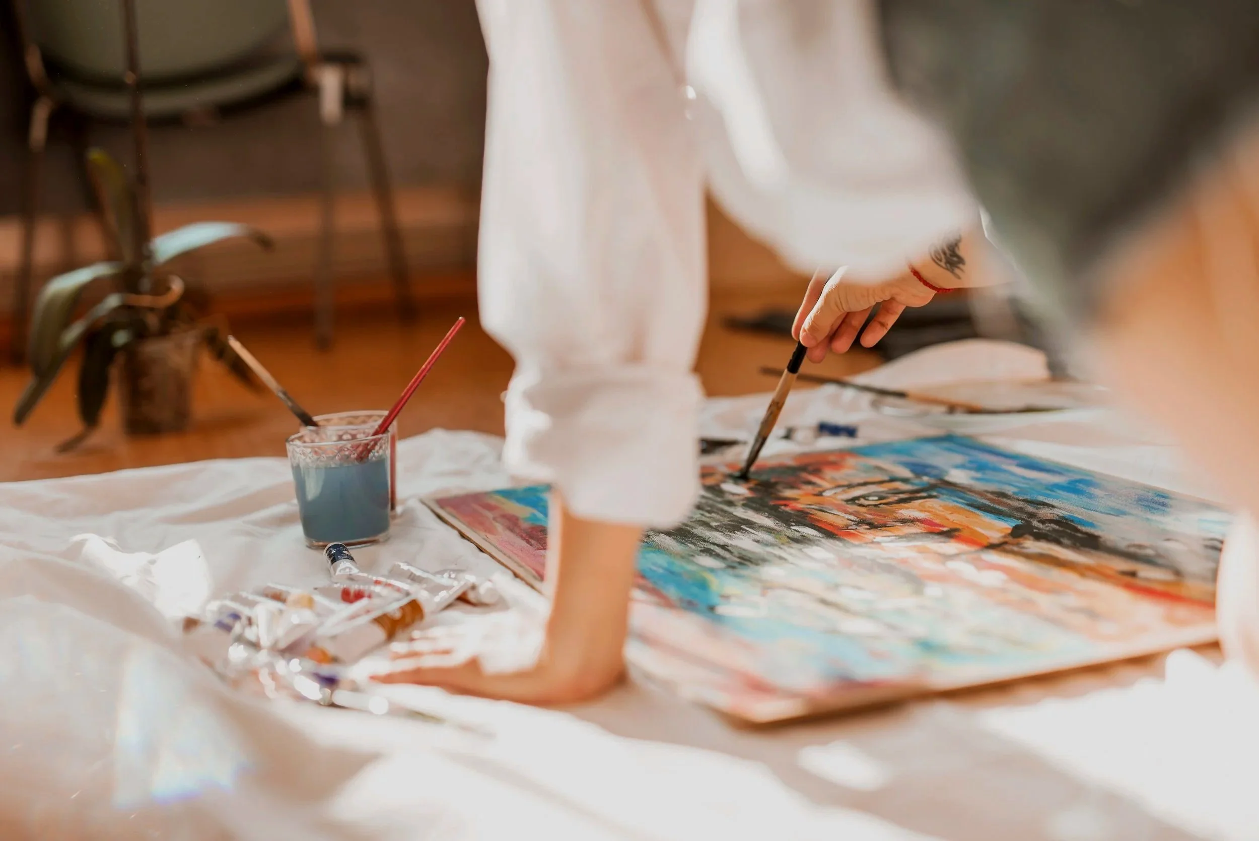 Person painting on a canvas with various paint supplies on a table, including tubes of paint, brushes, and a glass of water.