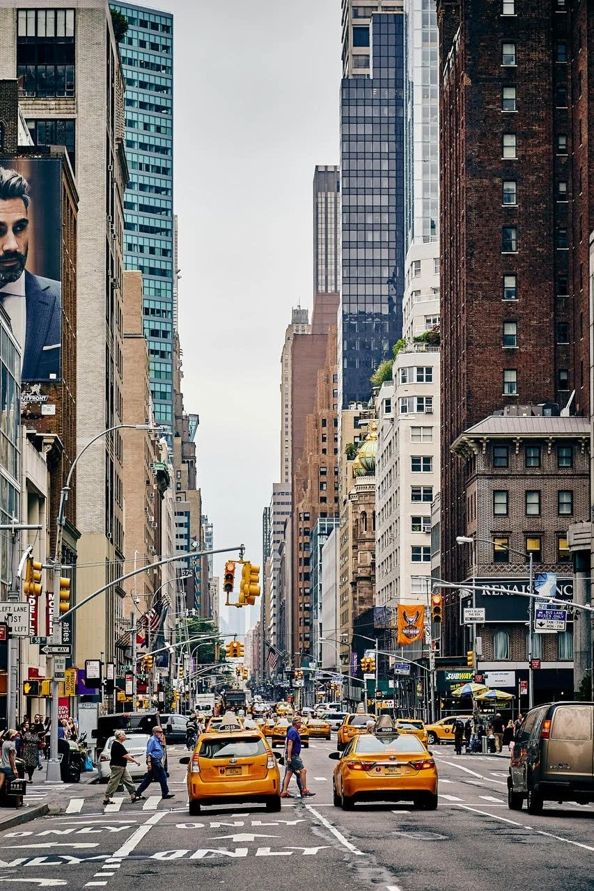 A busy city street scene with yellow taxis, pedestrians crossing, tall buildings on either side, and traffic lights