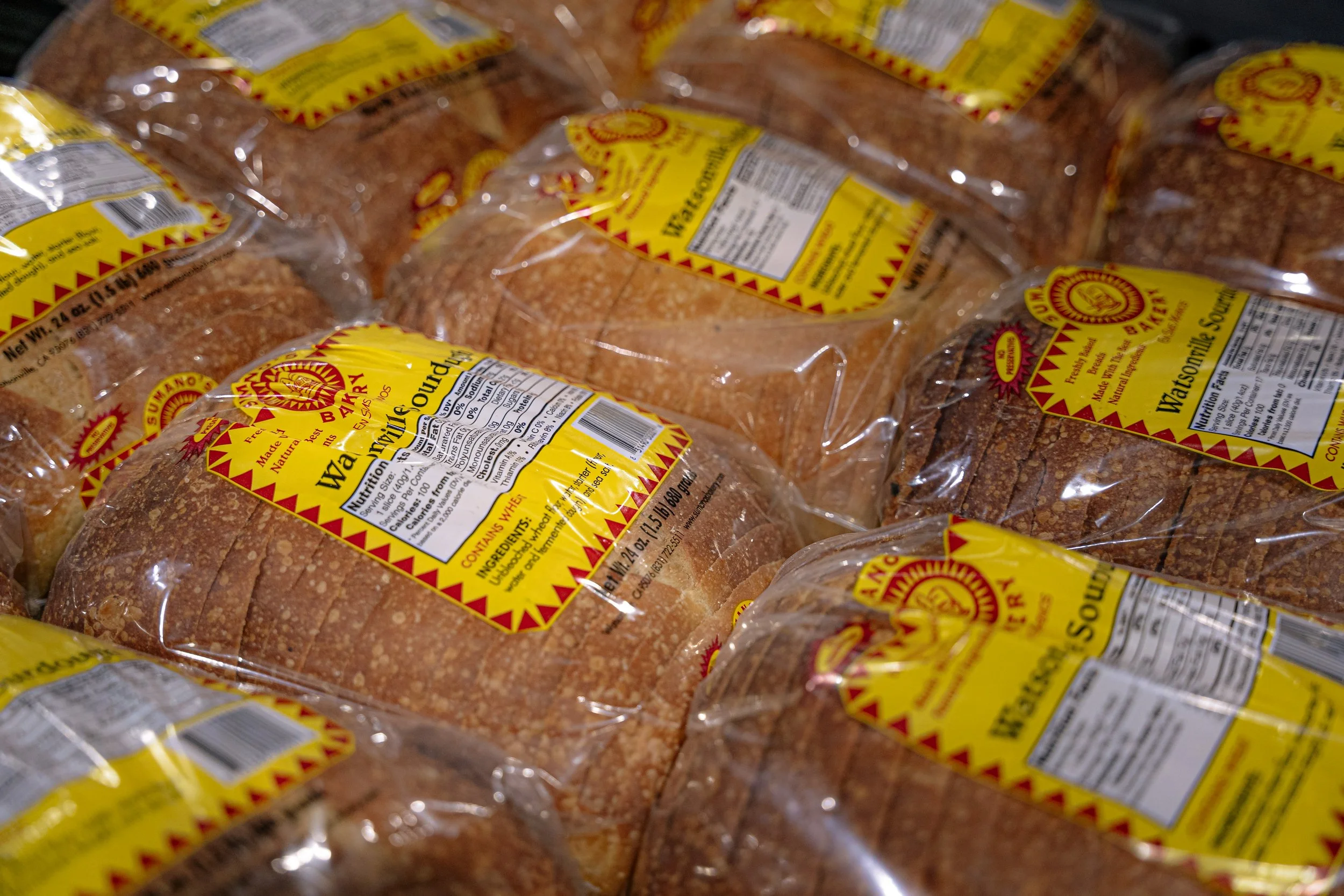 Multiple loaves of bread in plastic packaging with yellow and red labels, stacked on top of each other.
