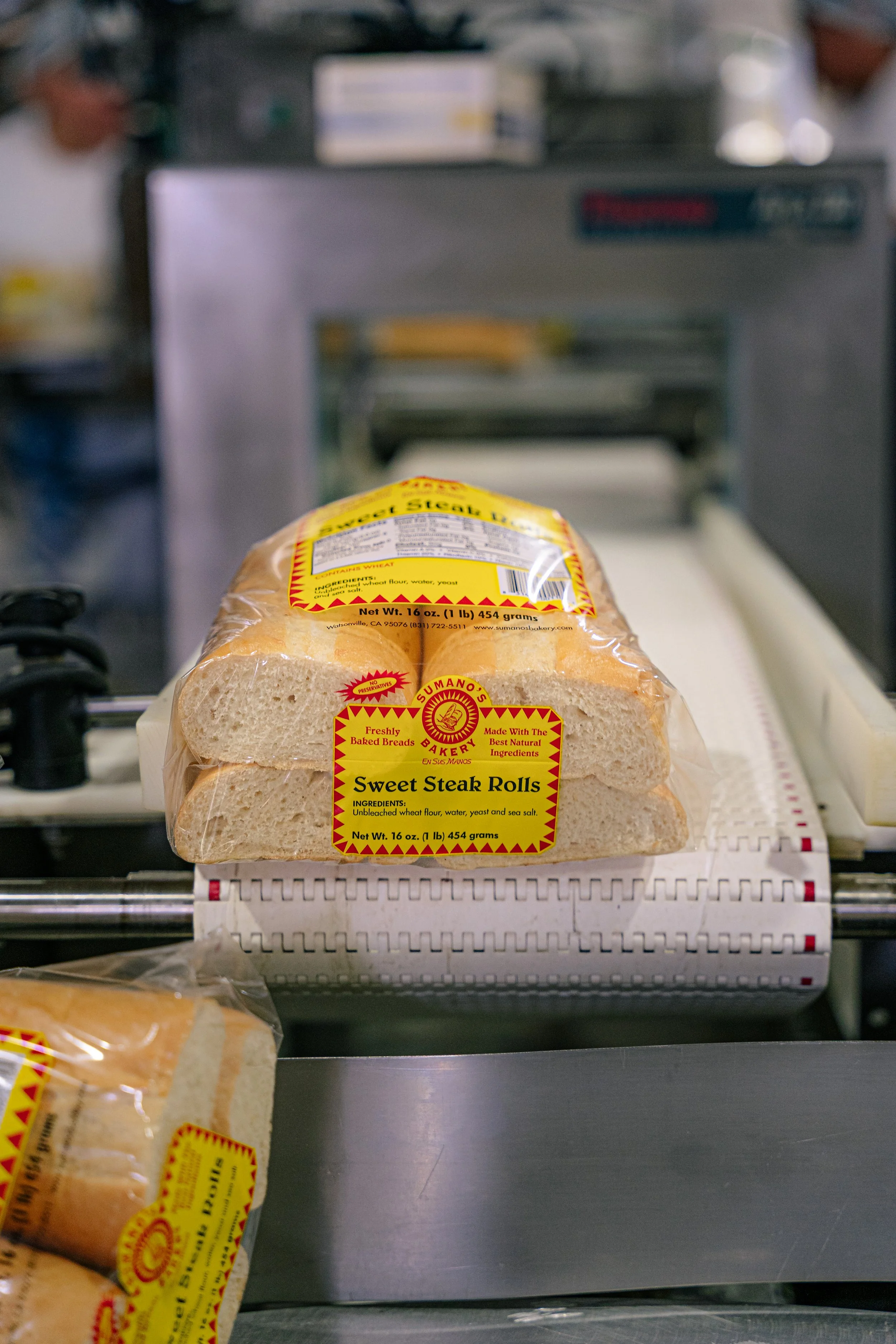 Loaf of Sweet Steak Rolls bread on a bakery conveyor belt, with blurred background of bakery equipment and staff.