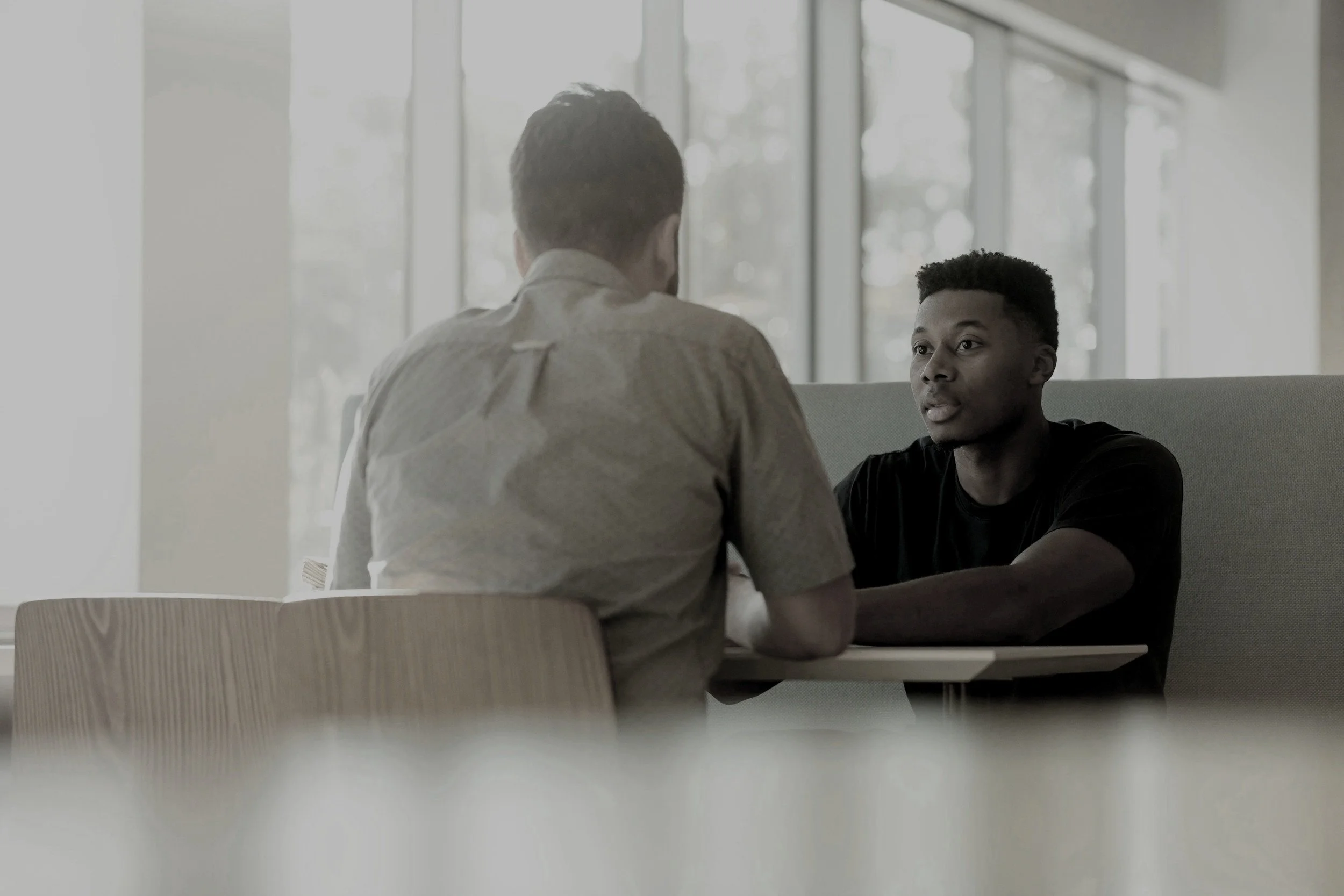 Two young men sitting across from each other at a table, engaged in a serious conversation, with a background of large windows letting in natural light.