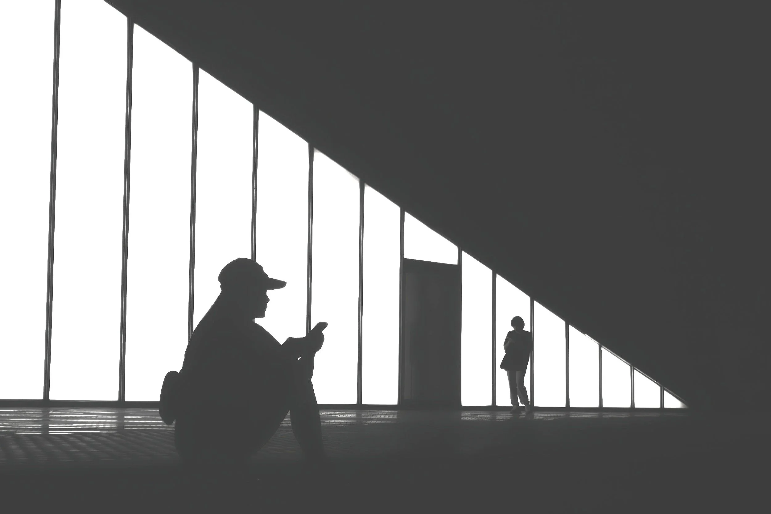 Silhouettes of two people, one sitting and one standing, against large windows in a modern building with an angled ceiling.