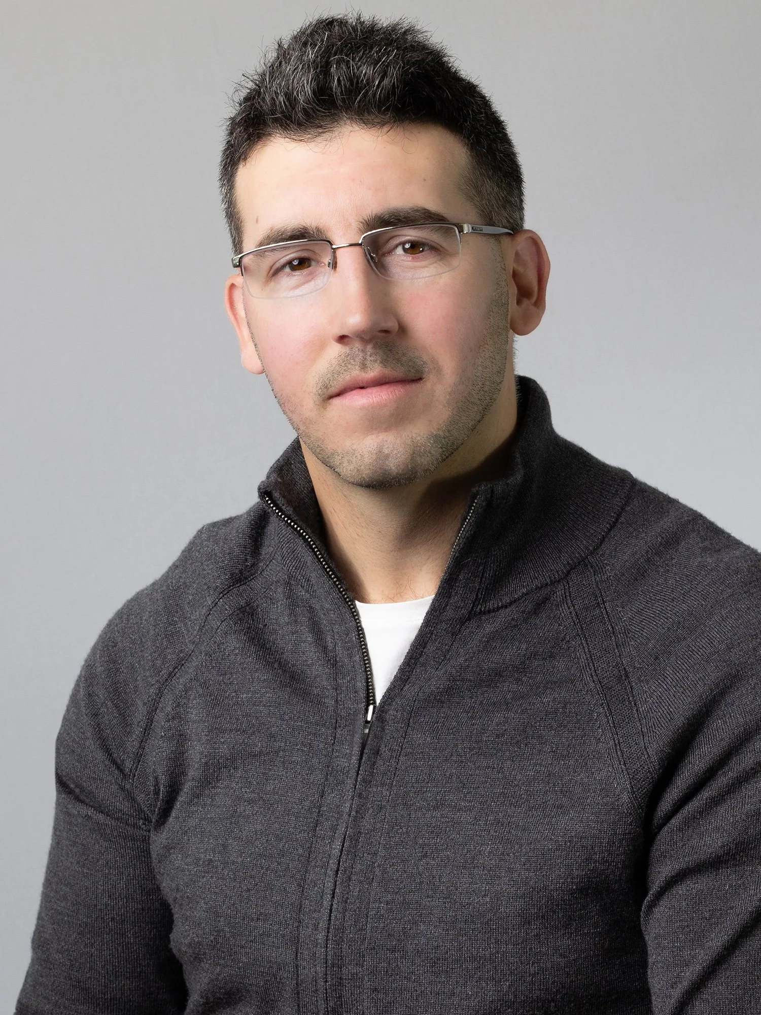 A man with short dark hair, wearing glasses and a black zip-up sweater, poses against a plain gray background.