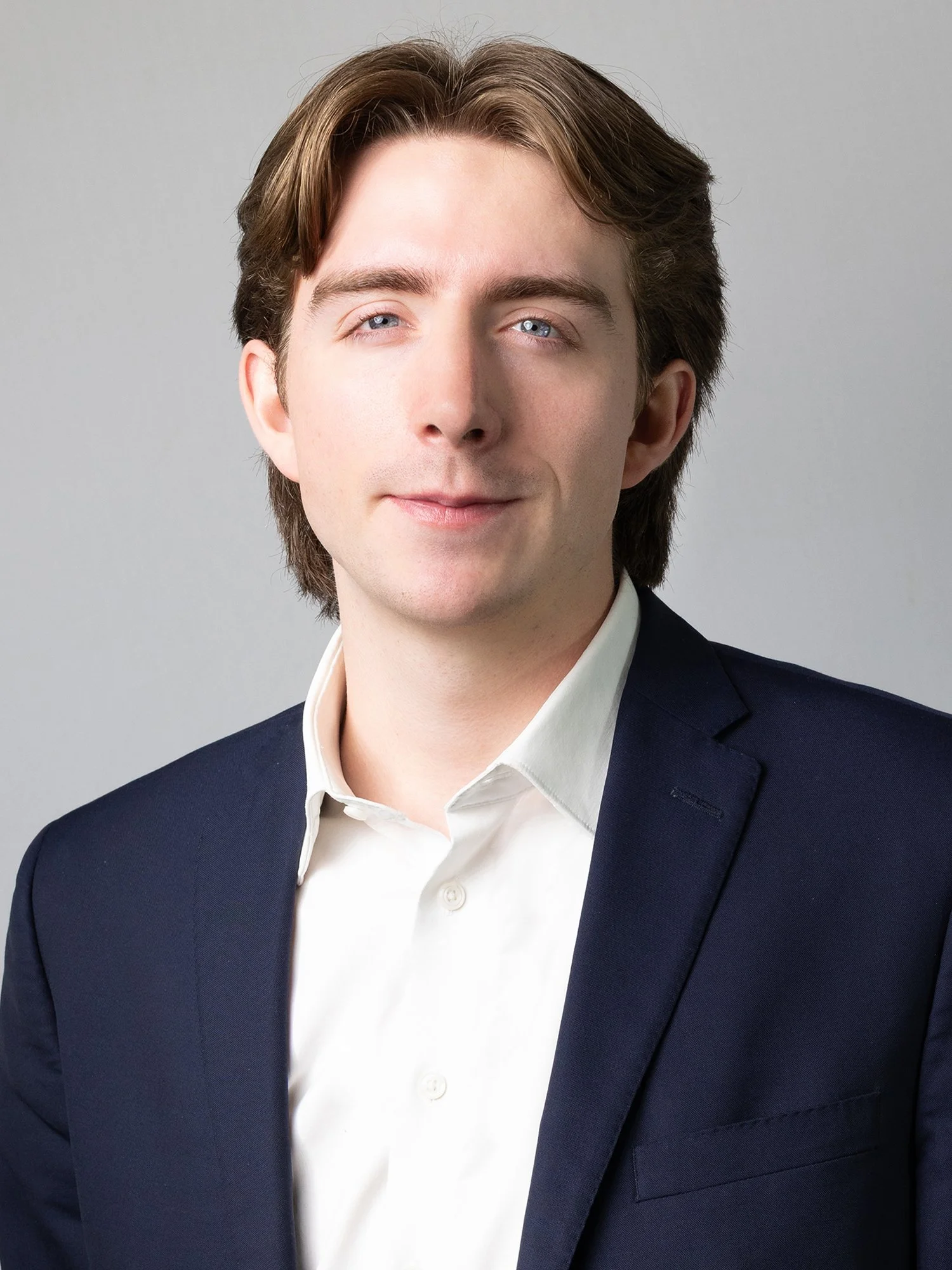 A young man with light skin, brown hair, and blue eyes is dressed in a white shirt and navy suit jacket, posing against a plain gray background.