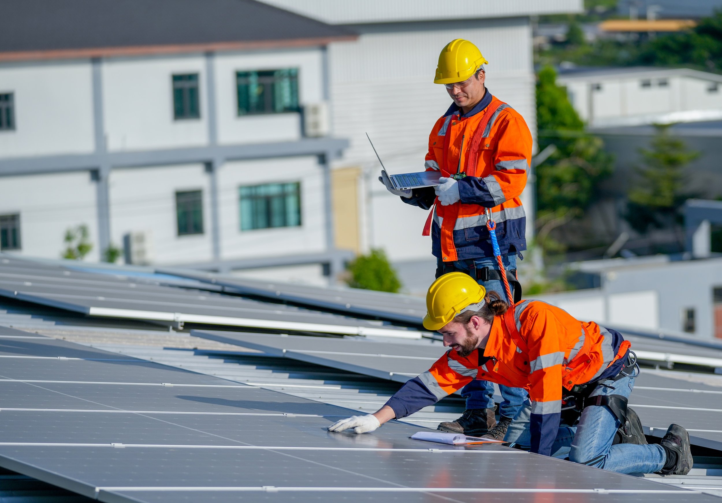 Two solar panel installers wearing orange safety jackets inspecting the panels while the other stands and looks at a laptop.