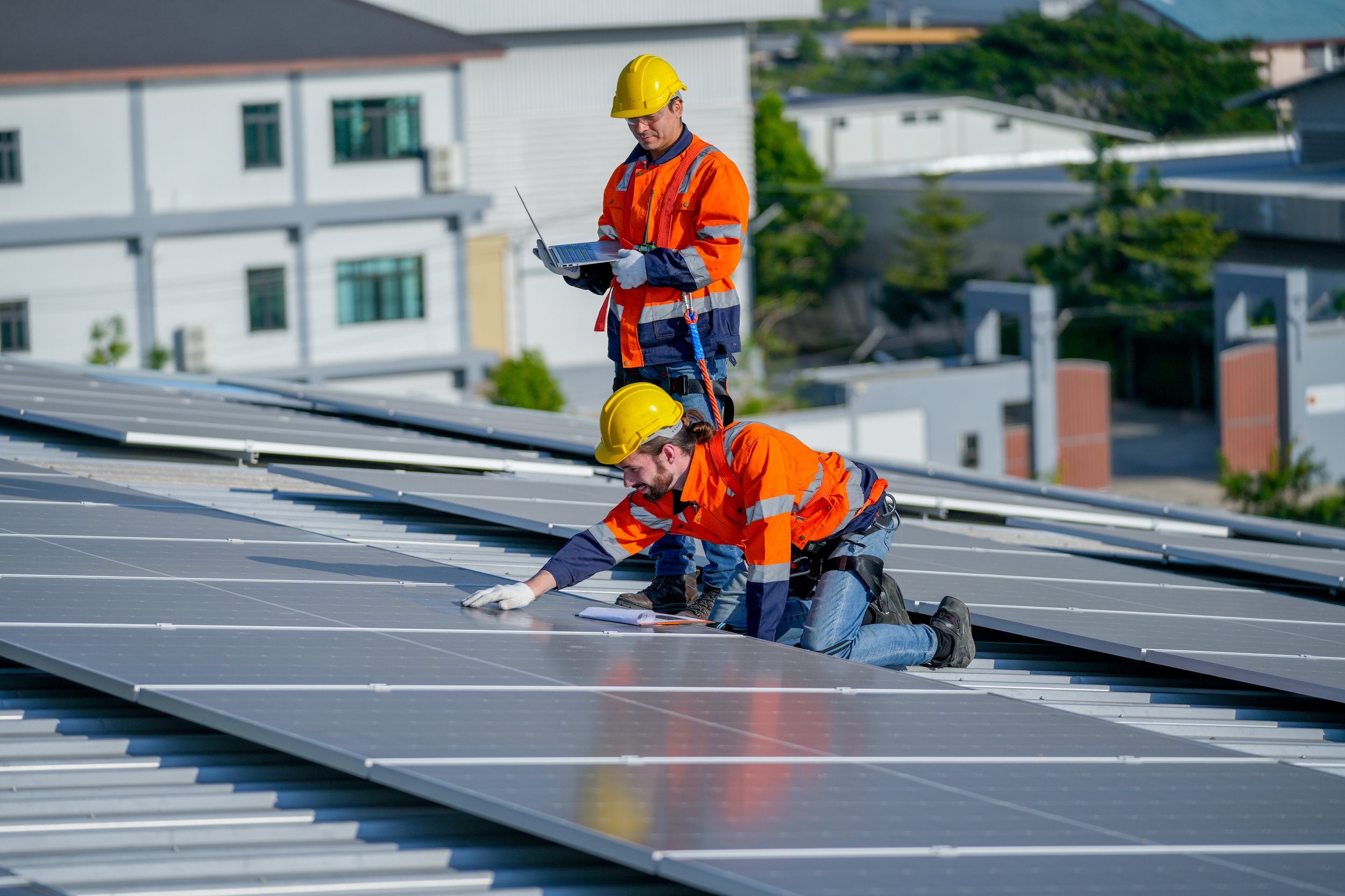 Two workers in orange safety jackets and yellow helmets installing or inspecting solar panels on a rooftop. One is kneeling and cleaning the panels, while the other stands holding a laptop.