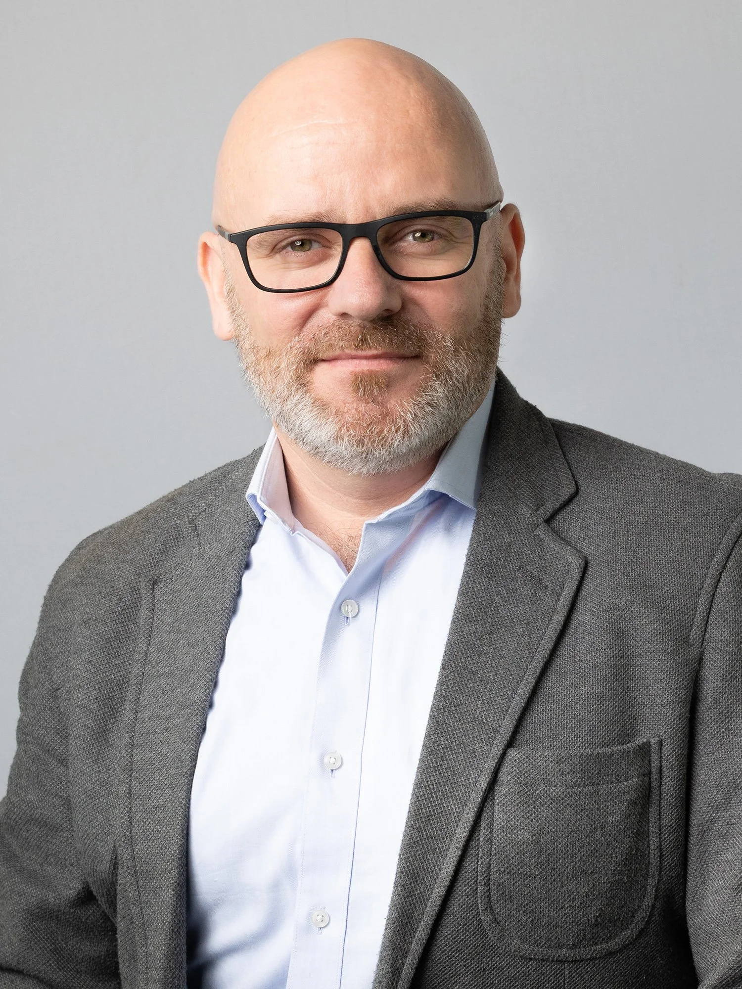 A professional man with glasses, a bald head, and a beard, wearing a gray blazer and a white shirt, standing in front of a neutral background.