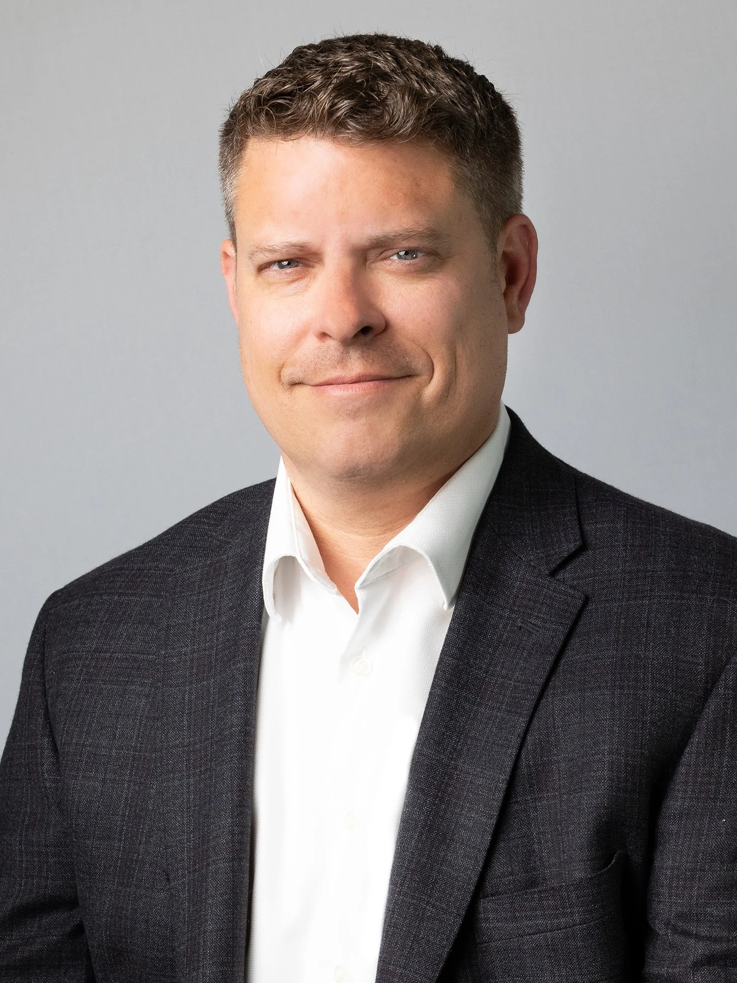 Headshot of a man in a white shirt and dark blazer smiling slightly against a plain gray background.