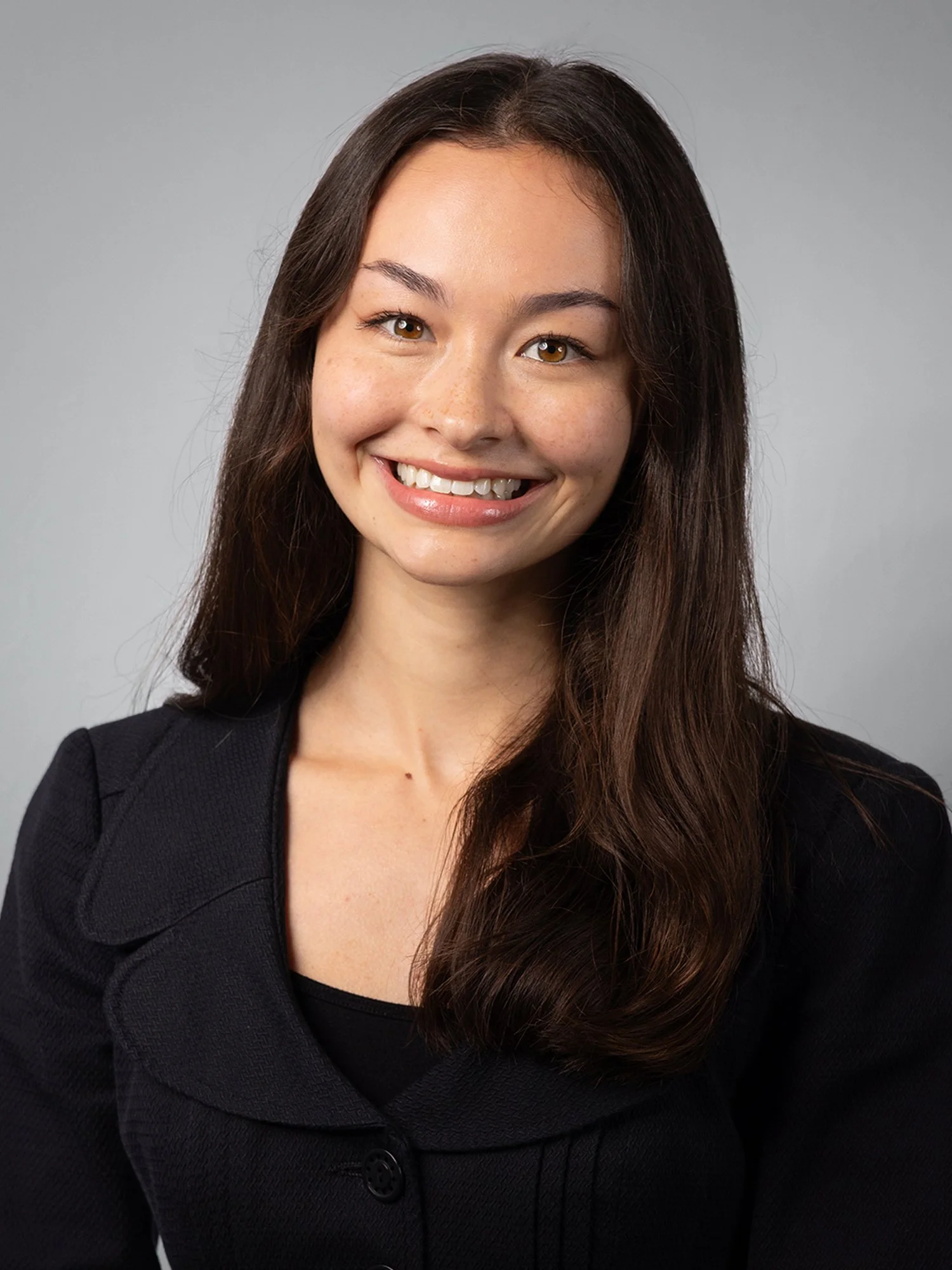 A woman with long brown hair, light skin, and freckles, wearing a black blazer and smiling at the camera against a plain light gray background.