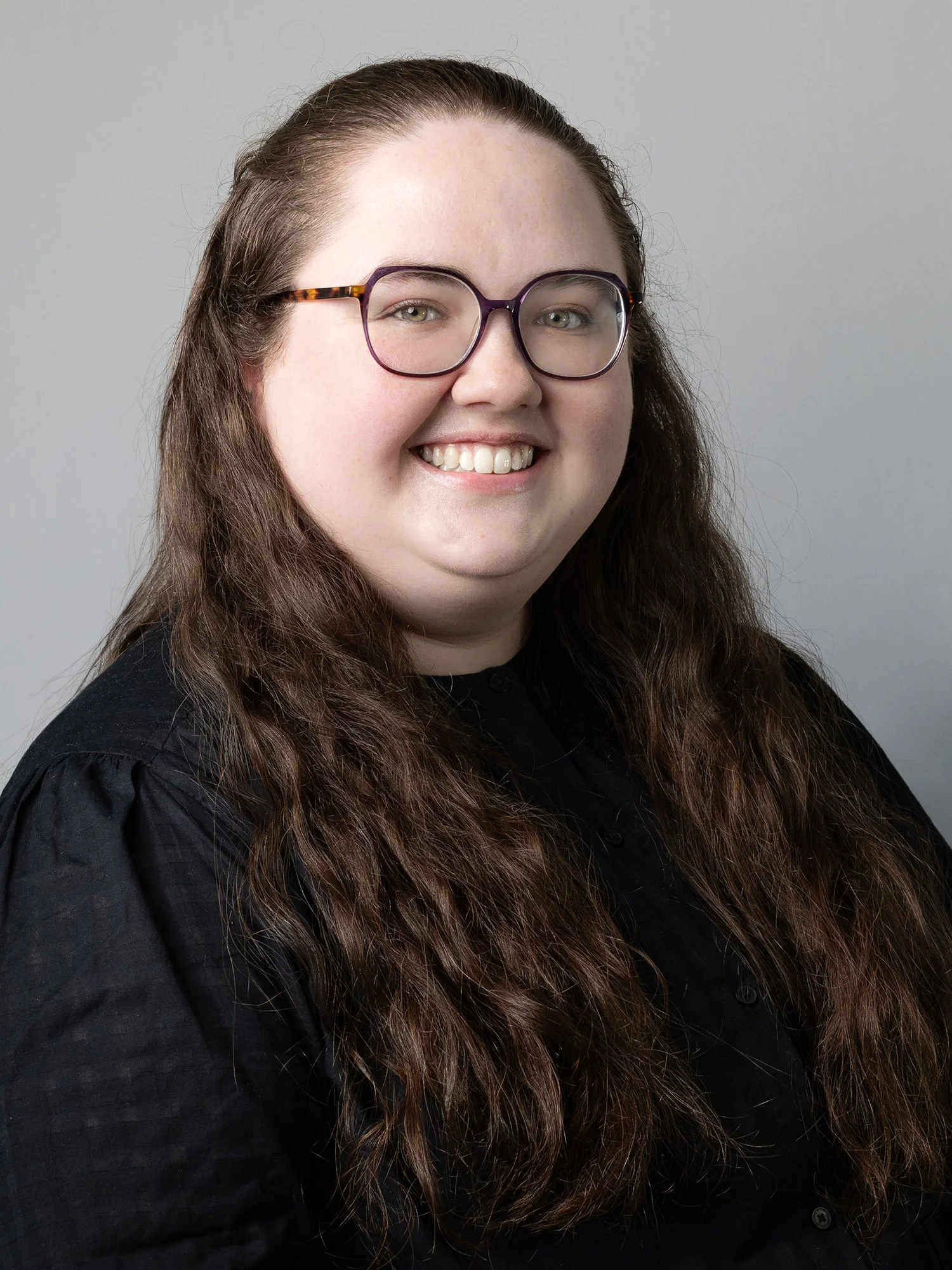 A woman with long, wavy brown hair wearing glasses and a black shirt, smiling at the camera against a gray background.