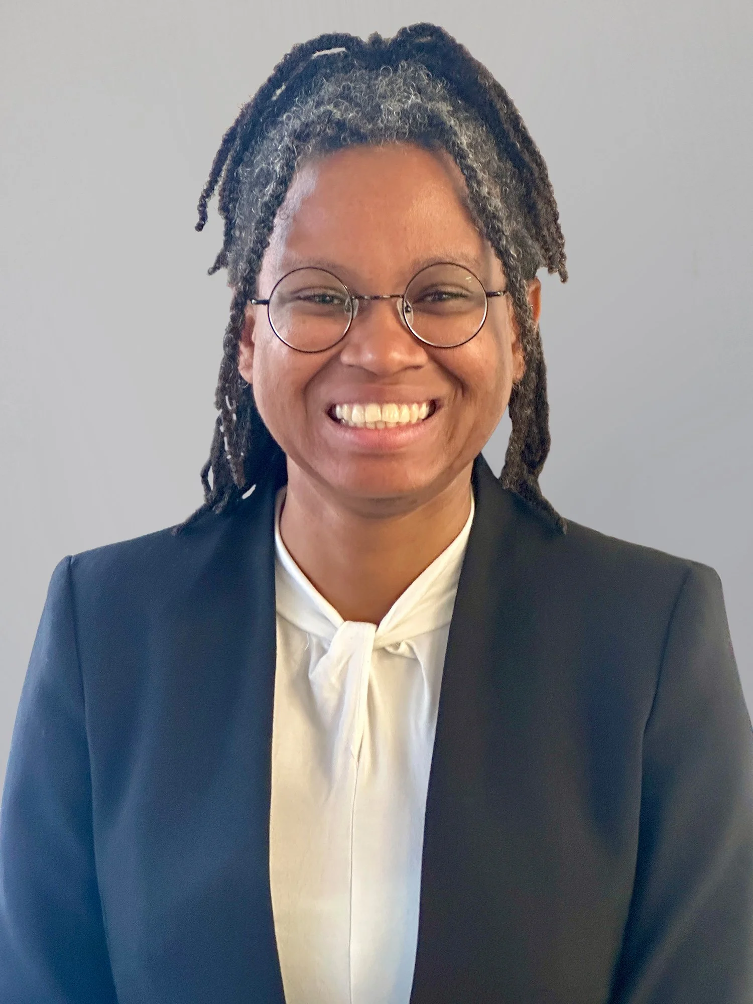 A smiling woman with glasses, wearing a dark blazer and a white blouse, standing against a plain grey background.