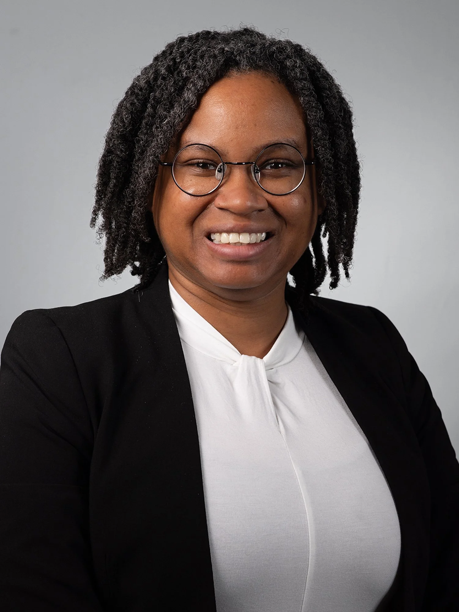A smiling woman with glasses, wearing a dark blazer and a white blouse, standing against a plain grey background.