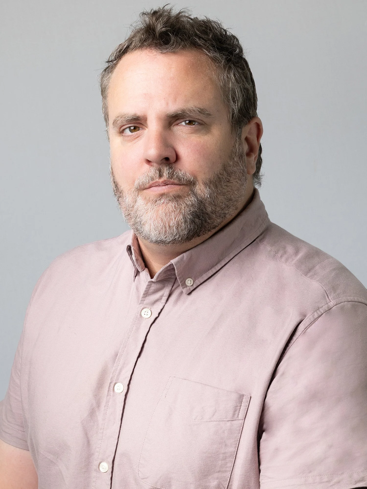 A middle-aged man with short gray hair and a beard, wearing a light pink button-up shirt, looking directly at the camera against a plain gray background.