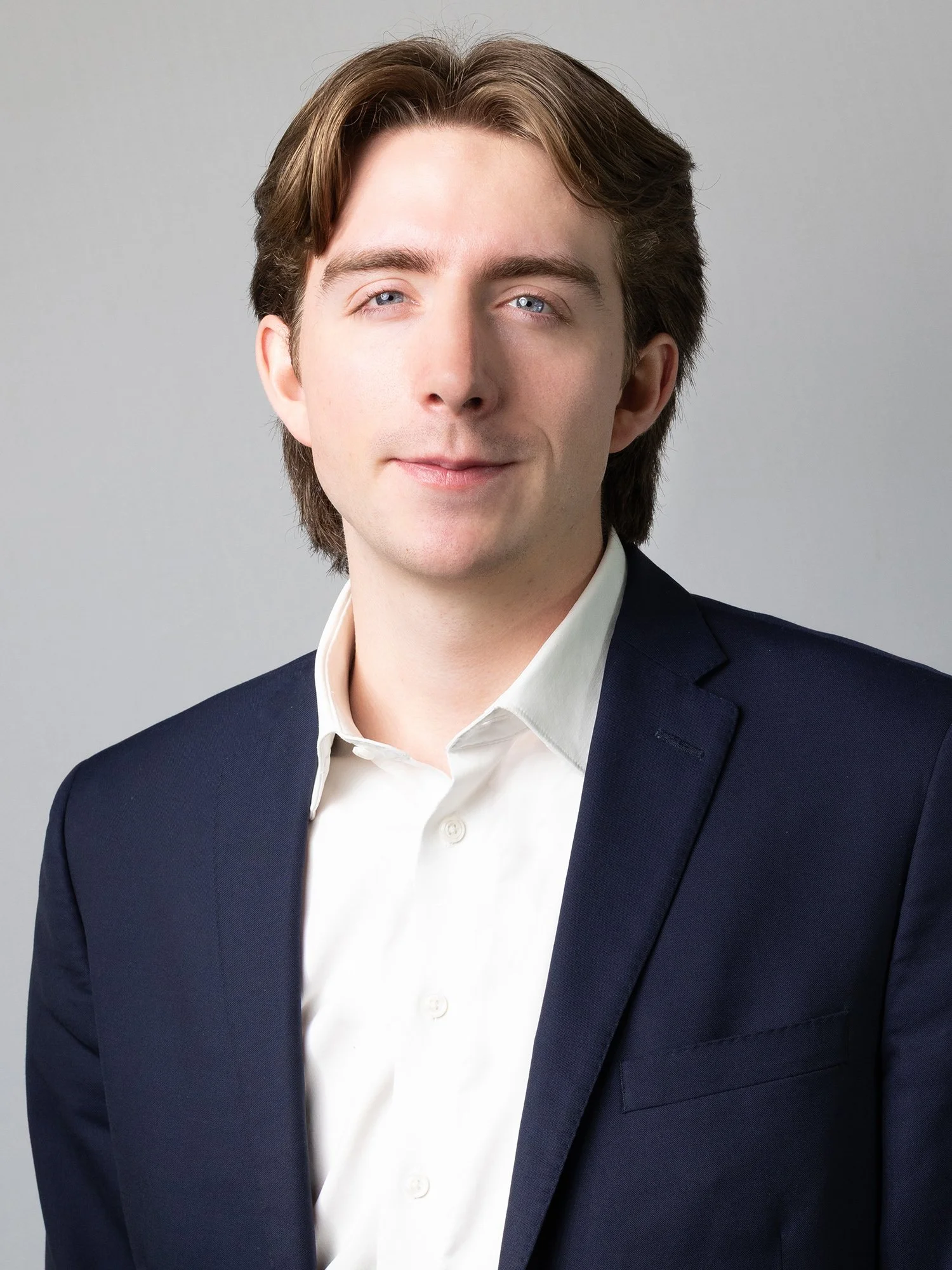 A young man with light skin, brown hair, and blue eyes is dressed in a white shirt and navy suit jacket, posing against a plain gray background.