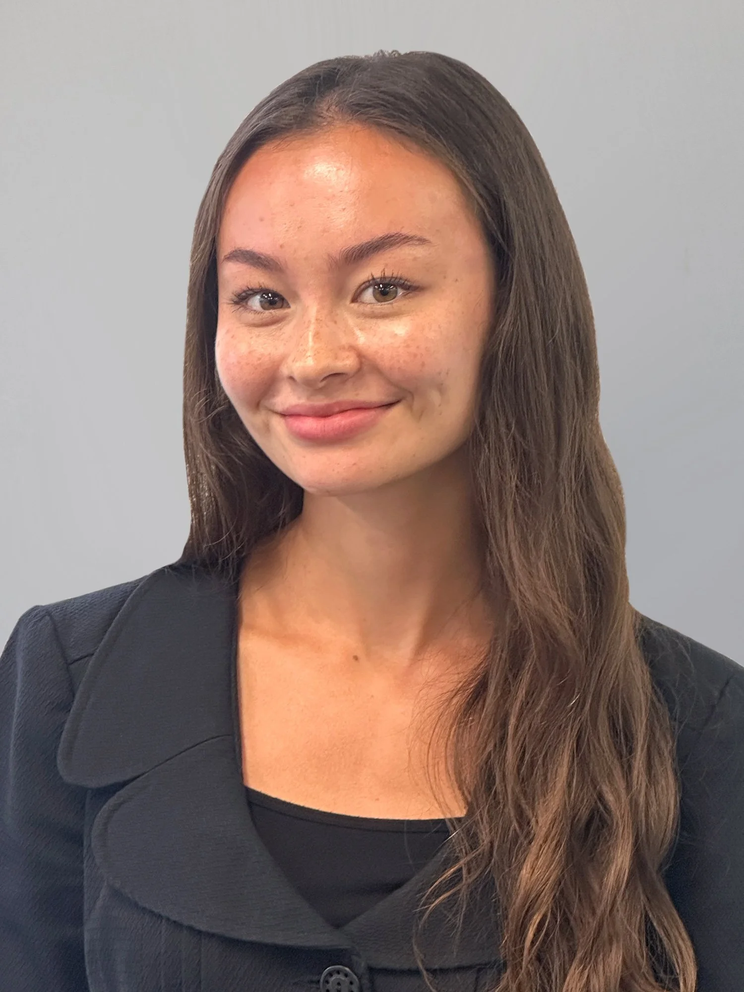 A woman with long brown hair, light skin, and freckles, wearing a black blazer and smiling at the camera against a plain light gray background.