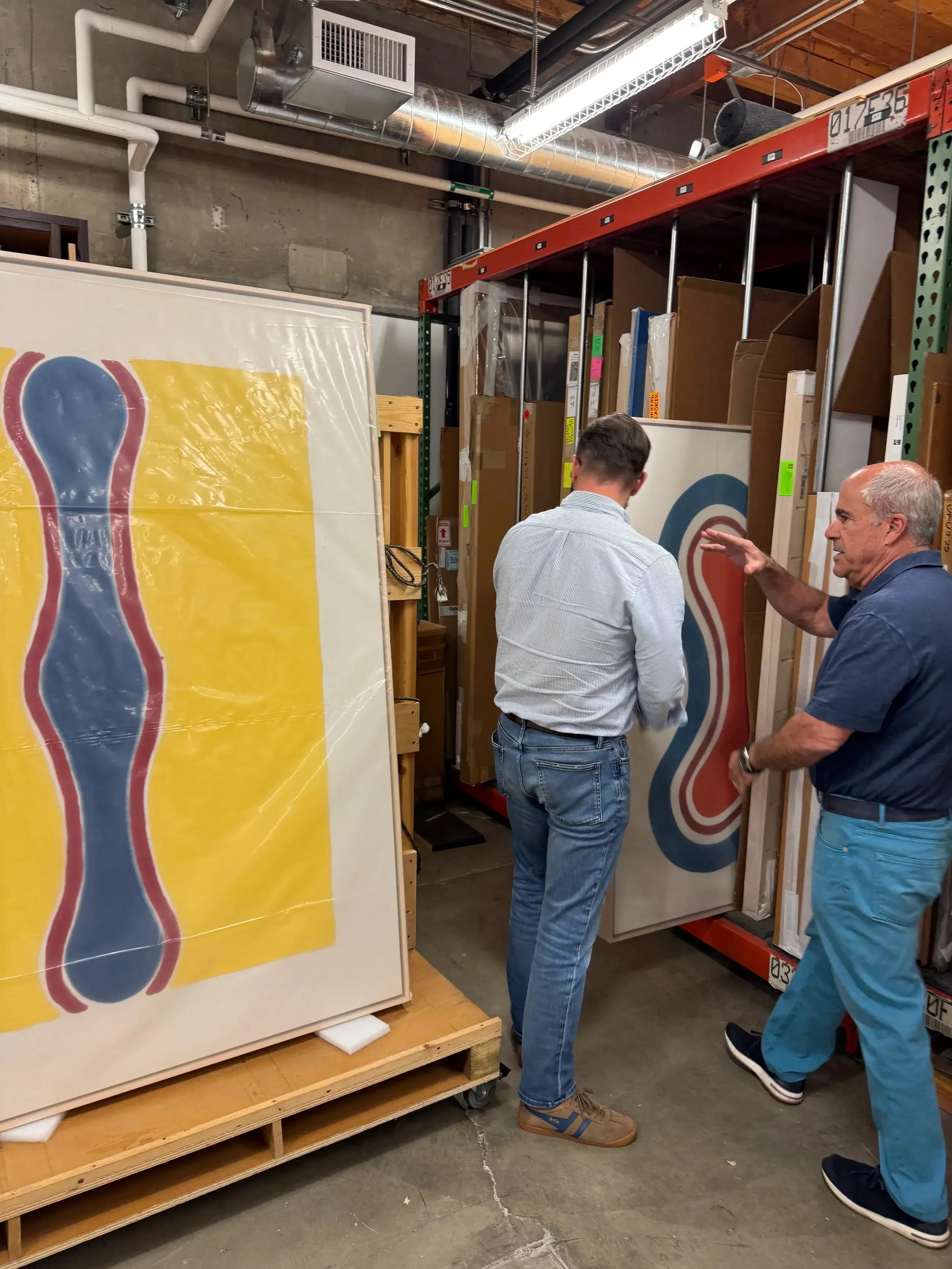 Two people stand in an art storage room discussing abstract paintings with wavy, colorful shapes. The scene conveys a professional, creative atmosphere.