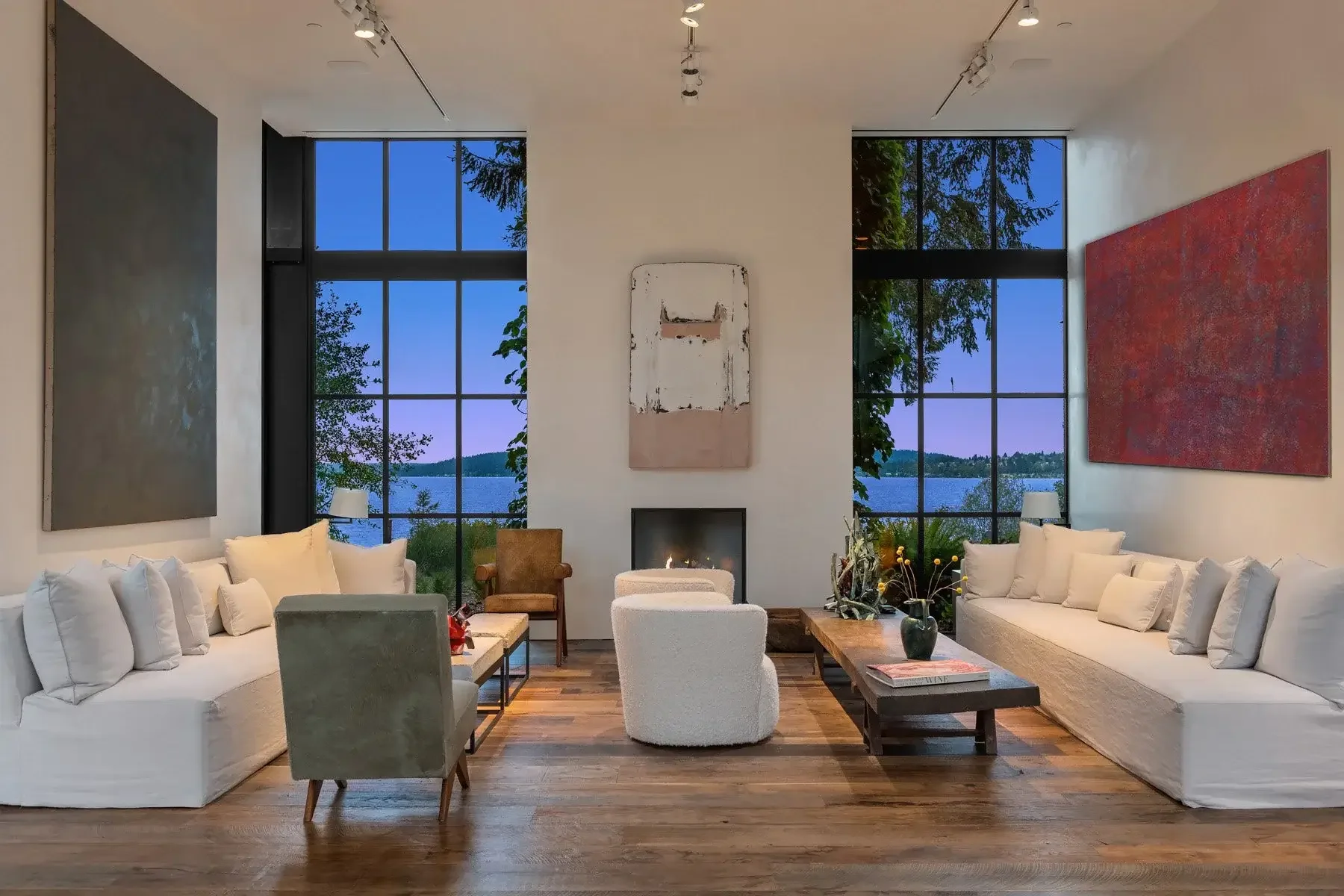 Spacious living room with large windows showcasing a lake view at dusk. Neutral sofas, abstract art, wooden floor, and a cozy fireplace create a serene atmosphere.