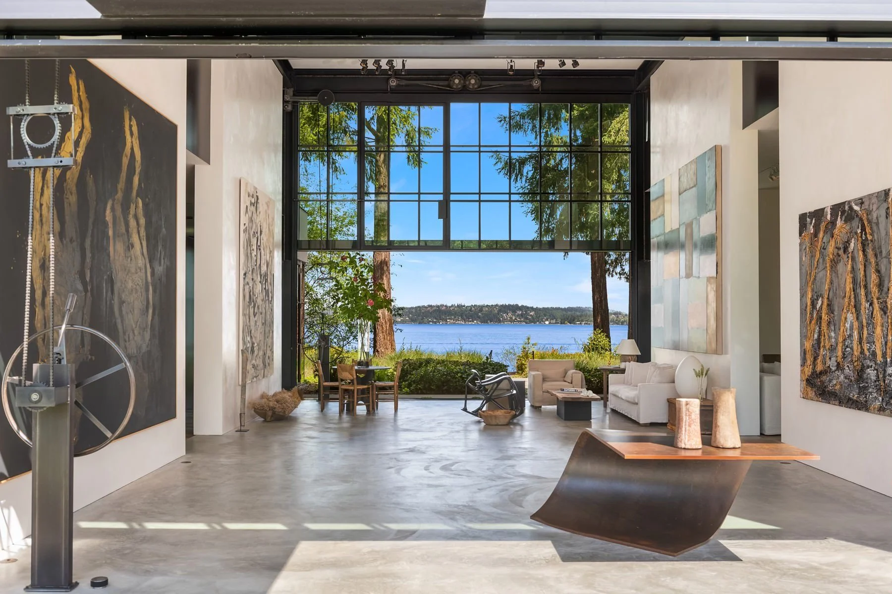 Modern industrial-style living space with high ceilings and large abstract paintings, featuring a massive floor-to-ceiling glass door that opens to a scenic lakefront view.