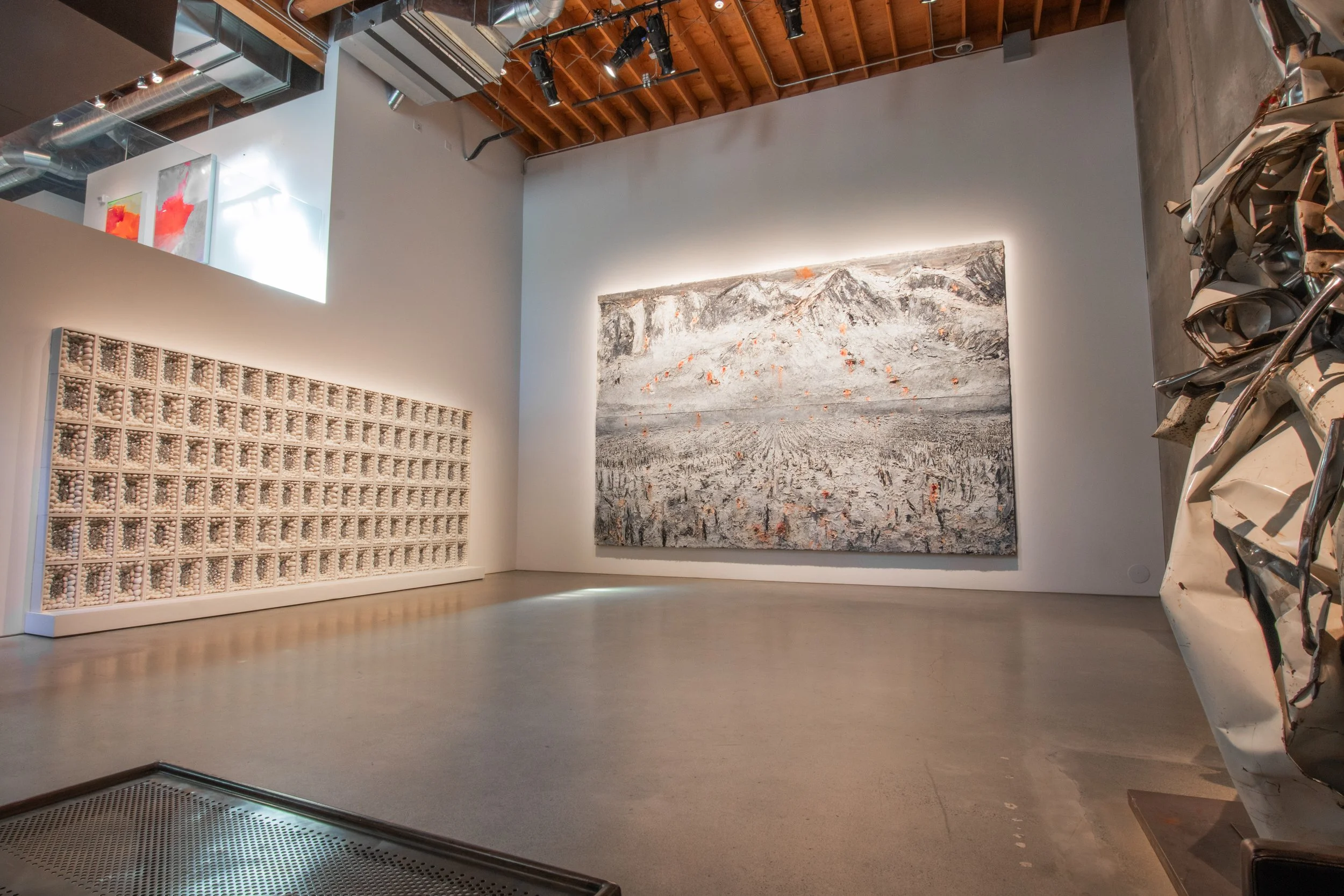 Spacious contemporary art gallery featuring a large-scale landscape painting, a textured grid wall installation, and a metallic sculpture in an industrial setting with exposed ceilings.