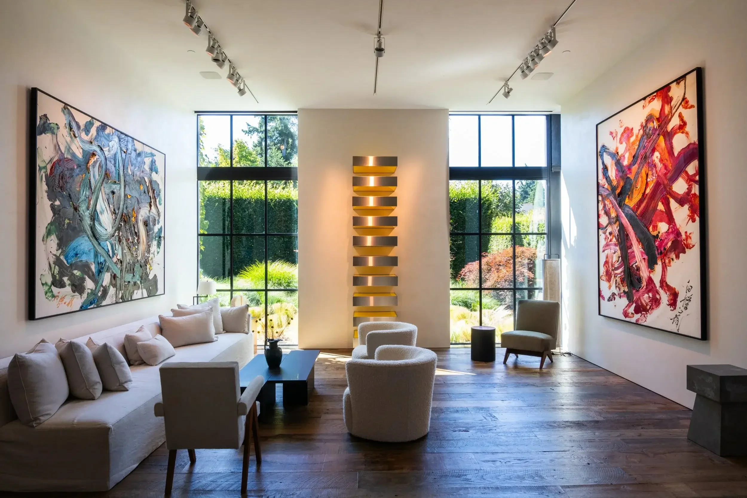 Modern living room with large abstract paintings on white walls, wood flooring, and floor-to-ceiling windows revealing a lush garden outside. Furniture includes a white sofa, cream armchairs, and a black coffee table.