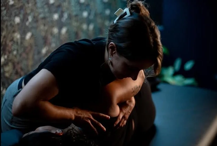 A woman leaning over a patient who is lying on their side on a therapy table, performing gentle osteopathy treatment on the shoulder for their shoulder pain and back pain.