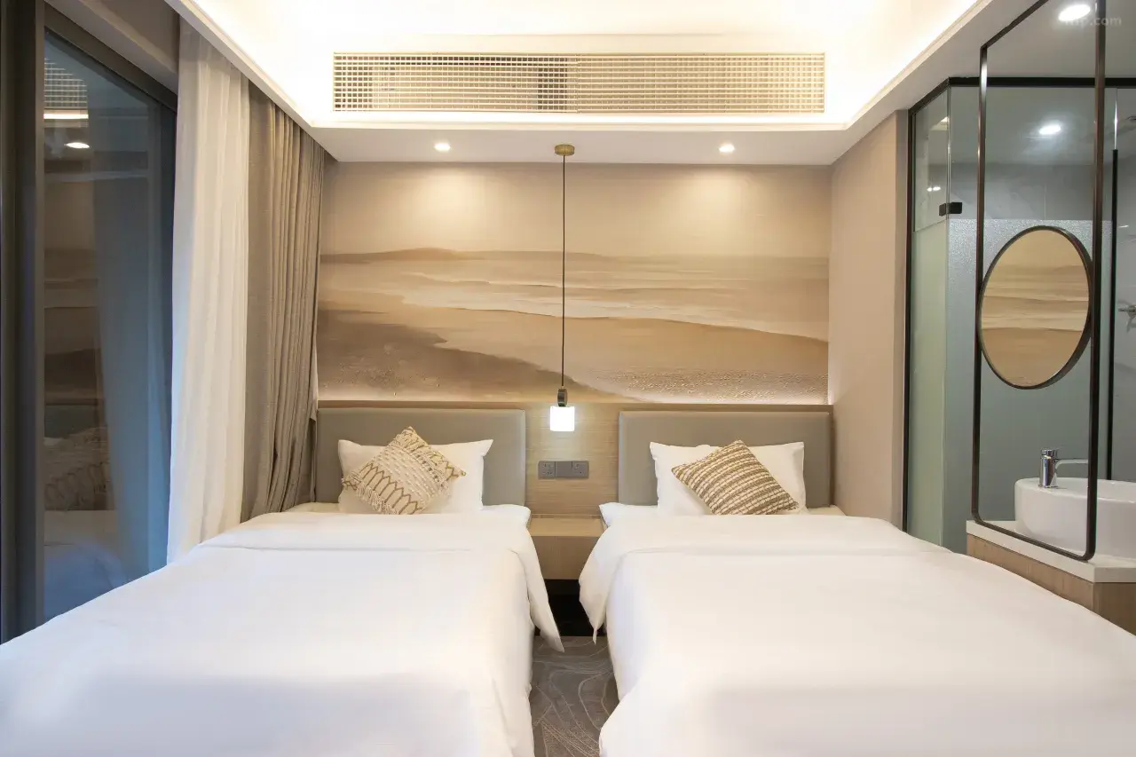 Hotel room with two single beds, decorative pillows, a wall mural of a beach scene behind headboards, and a glass-walled bathroom with a sink inside.