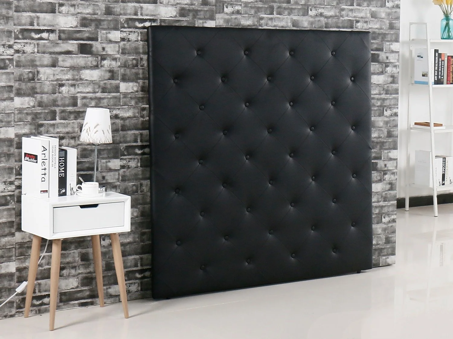Modern bedroom with a black padded headboard, gray brick wall, white nightstand with a drawer, books, a lamp, and a white tiered bookshelf with books and decorative items.
